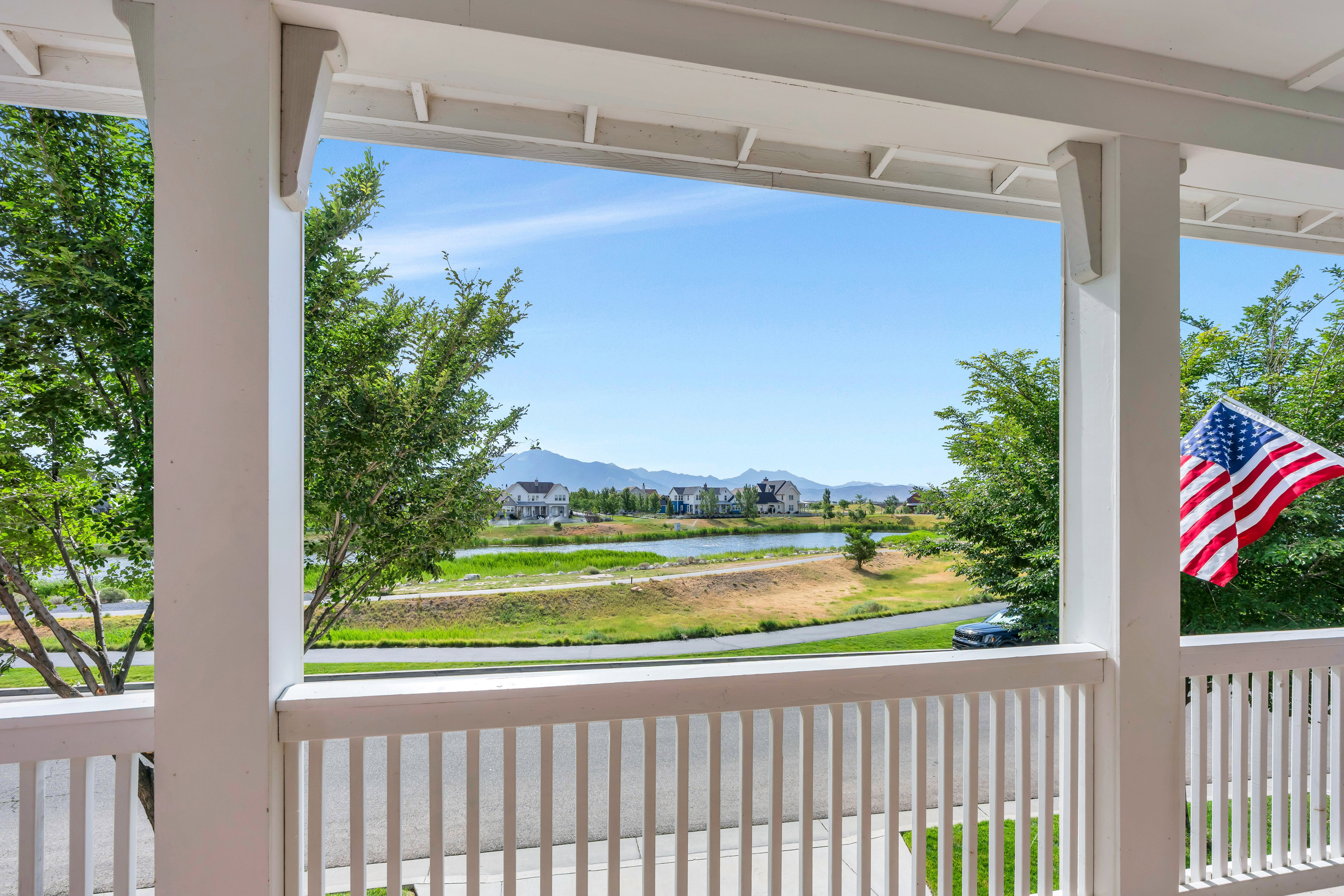 Front porch with white columns and railing, providing a direct sightline to a community pond, distant mountains, and residential structures beyond