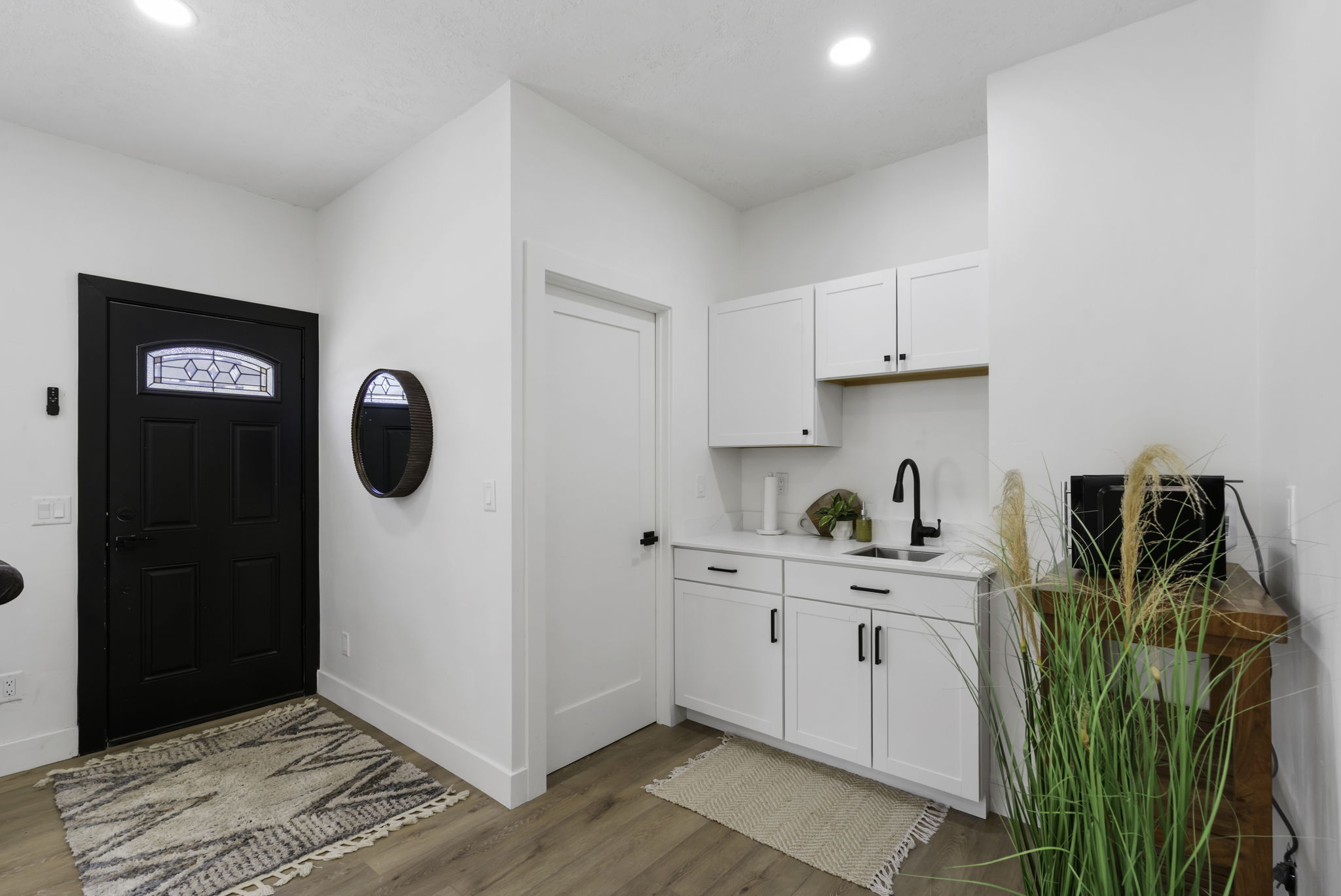Entryway featuring a black paneled door with decorative glass, wood-finish flooring, white walls, recessed lighting, and a compact kitchenette with white cabinetry, a black sink, and a black faucet