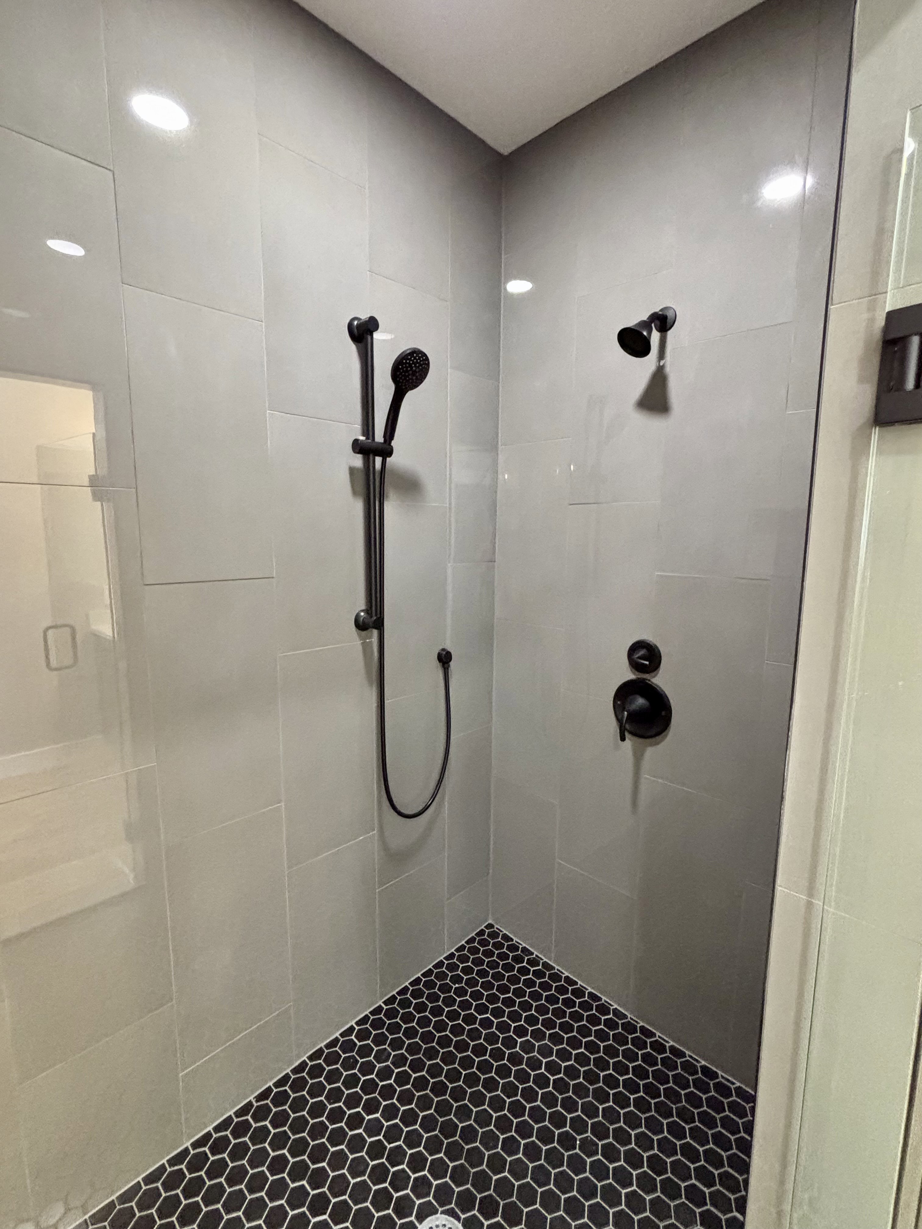 Modern shower enclosure featuring gray rectangular wall tiles, dark hexagonal floor tiles, a handheld shower fixture, a fixed shower head, and matte black hardware