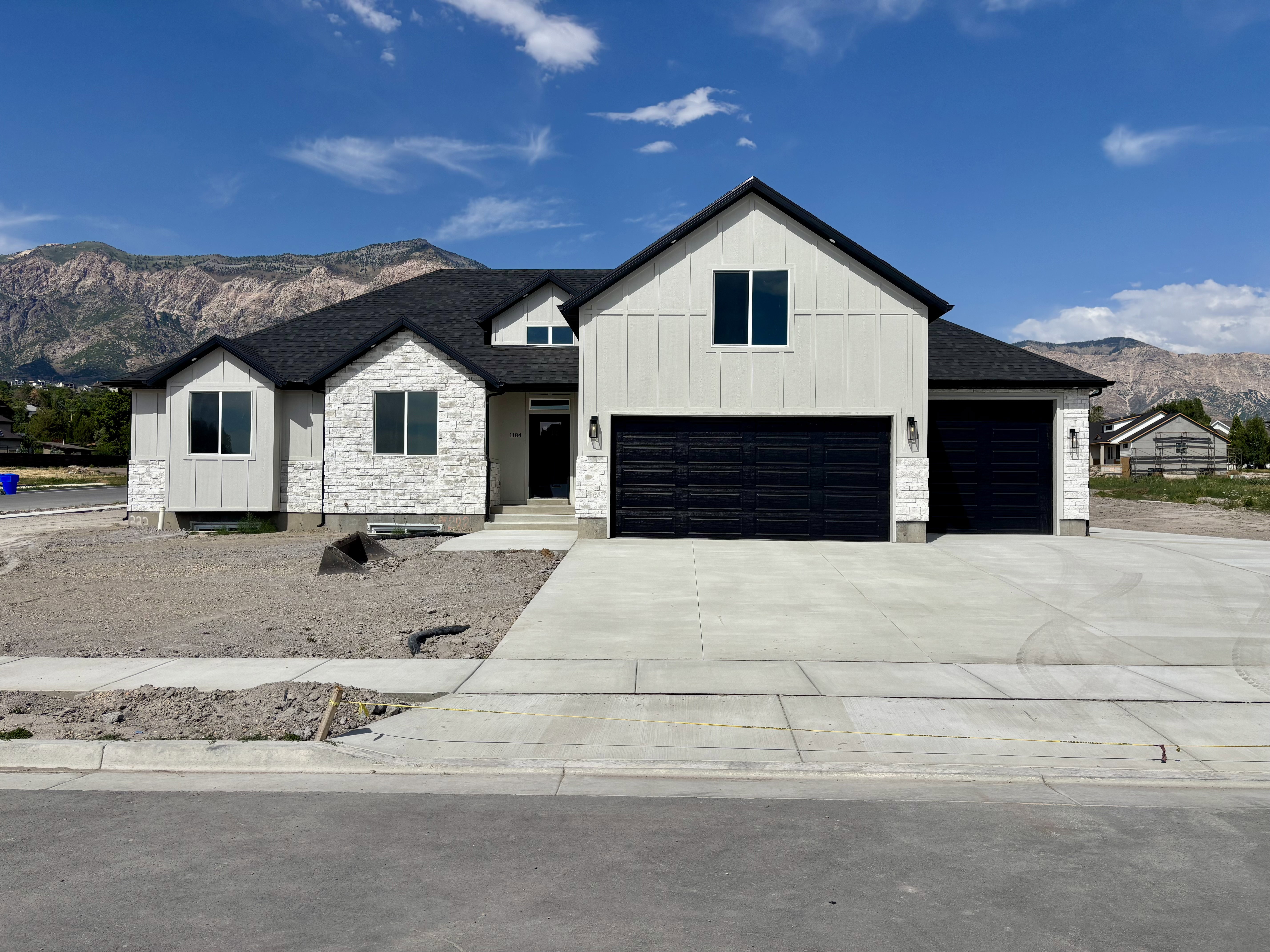 Modern farmhouse exterior featuring vertical white siding, white stone accents, a dark shingle roof, two black garage doors, and a concrete driveway