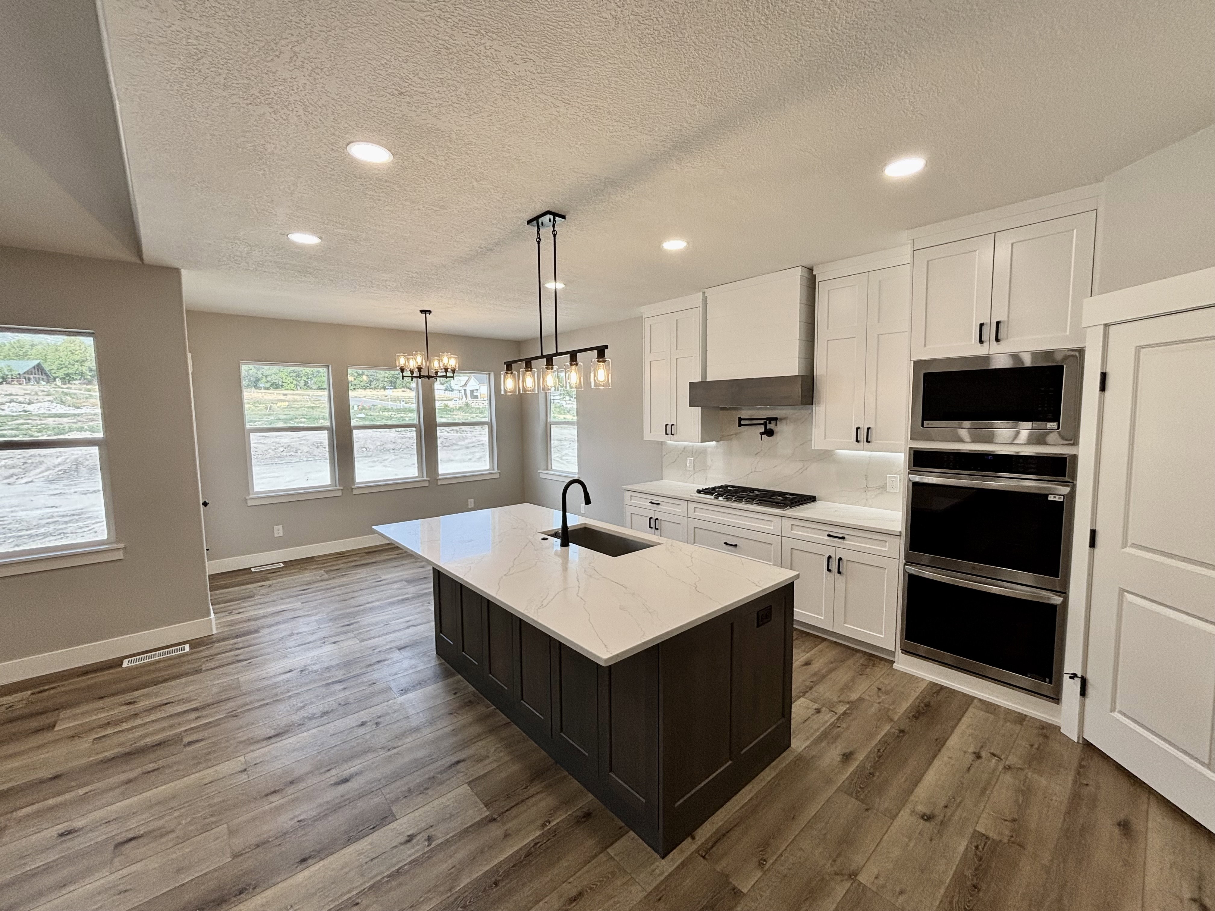 Spacious open-concept kitchen featuring a dark-toned island with a white countertop and integrated sink, light wood-finish flooring, white cabinetry with black hardware, and stainless steel double ovens