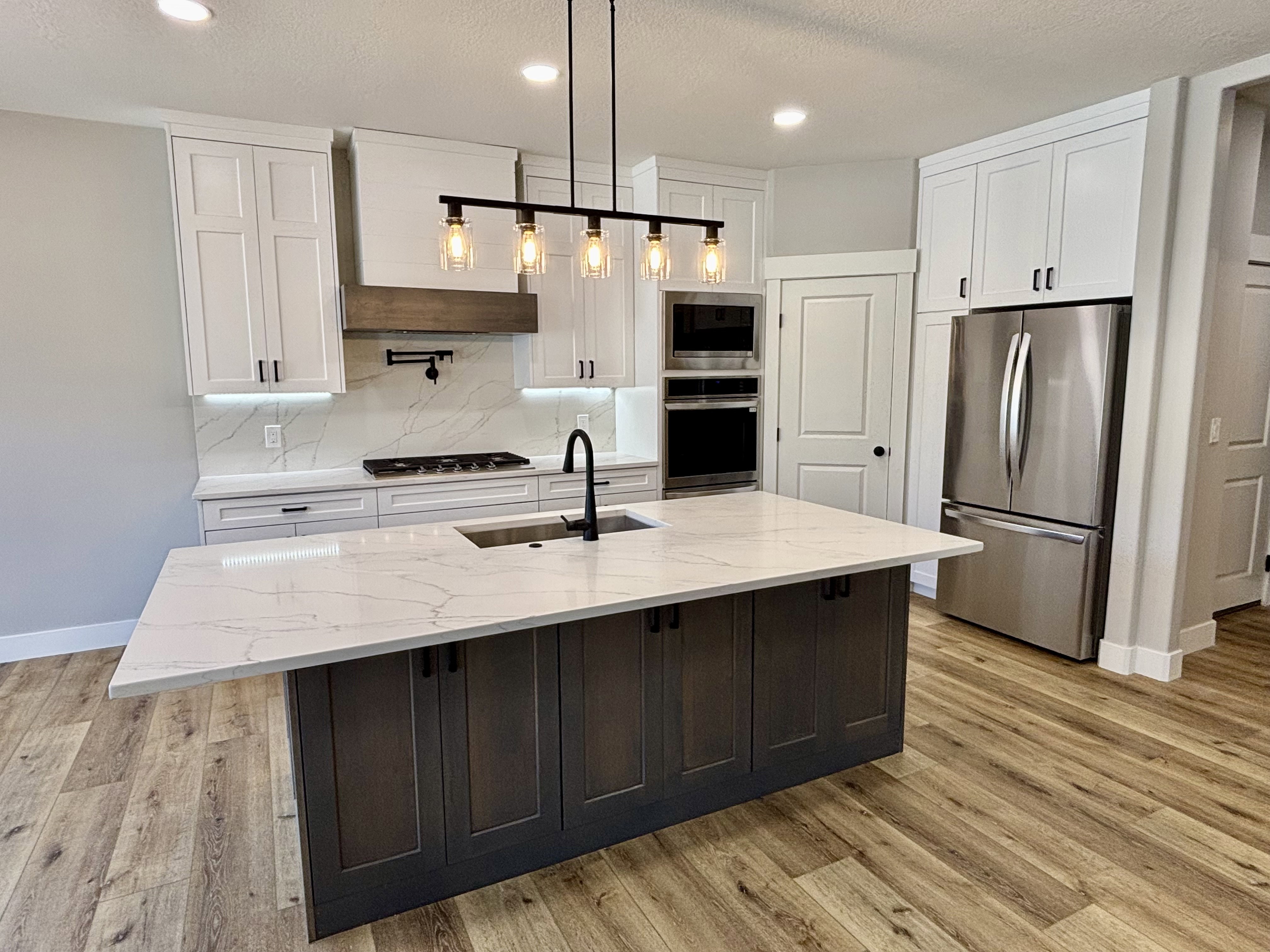 Kitchen featuring a central island with a light-toned quartz countertop, white cabinetry, integrated stainless steel appliances, gas range top, lvp wood style  flooring, recessed lighting and kitchen island pendant lighting