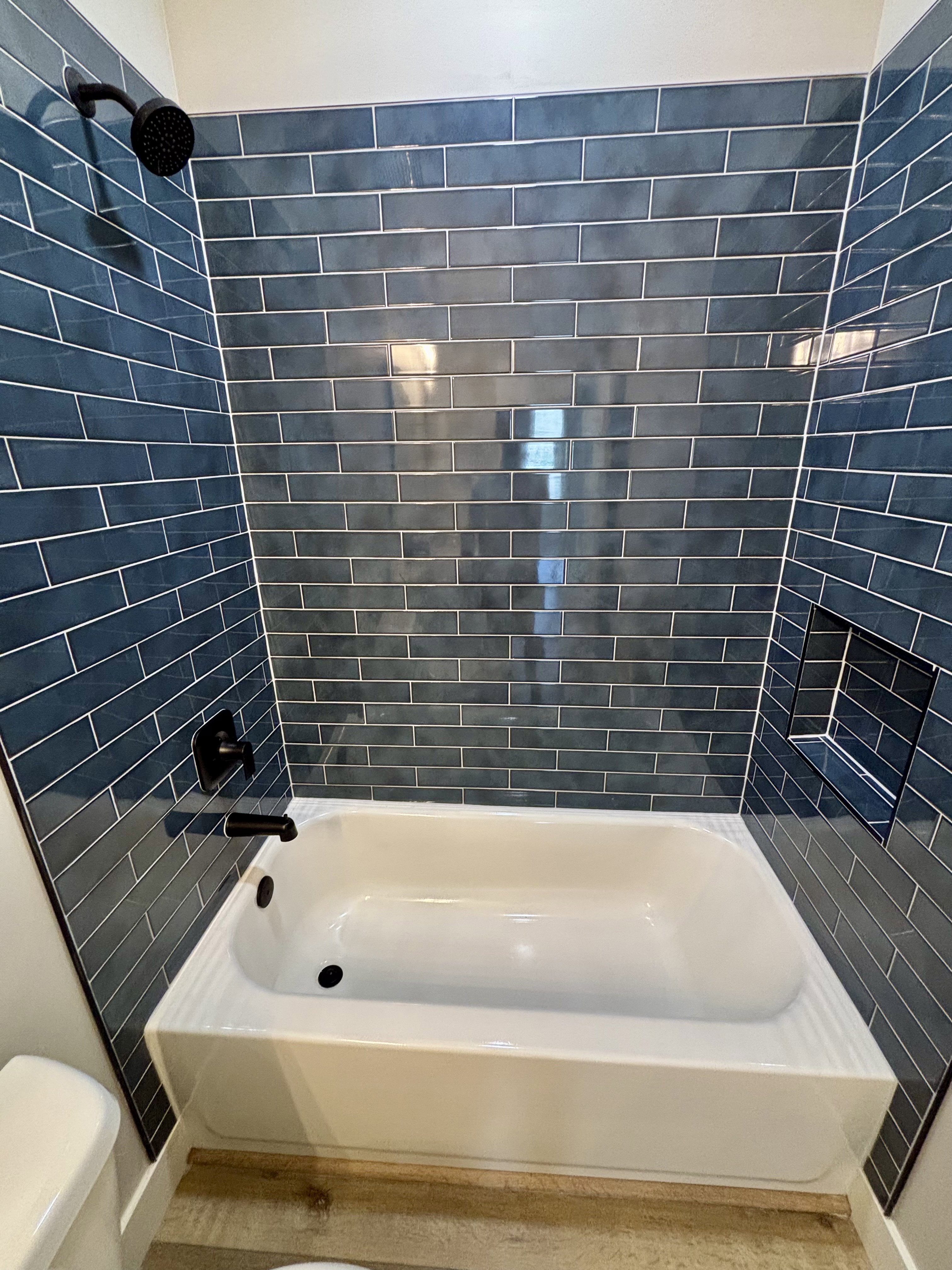 Bathroom featuring a white tub, dark blue subway tile shower surround, matte black shower fixtures, and a recessed shower niche