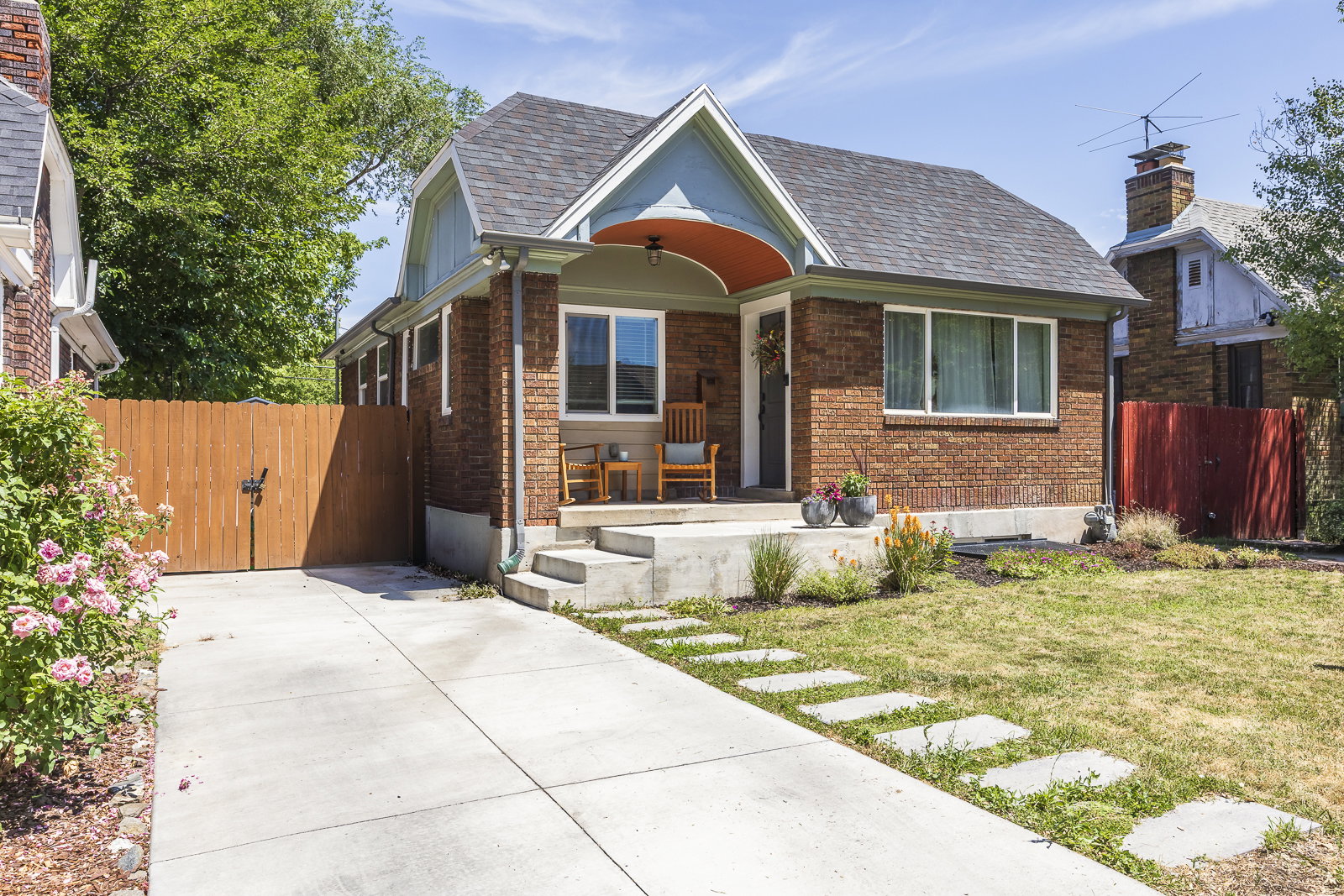 Brick facade residence featuring a covered porch with an arched entry, concrete front steps, a concrete driveway, a wooden privacy fence, and a front yard with stepping stones