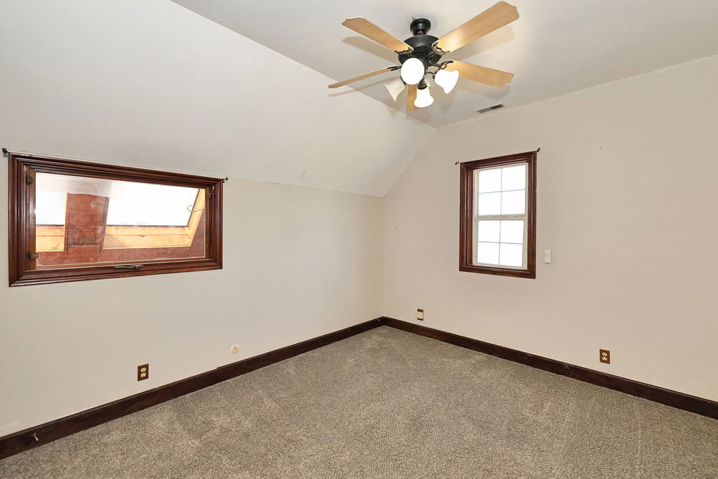 Carpeted room featuring a ceiling fan with light fixture, two distinct windows with dark wood trim, and sloped ceiling details