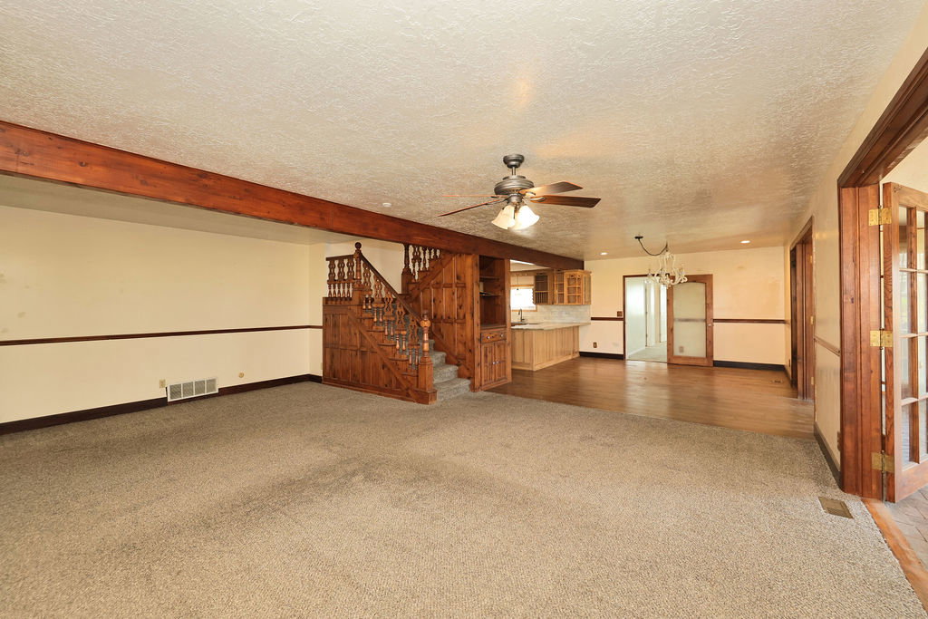 Carpeted living area featuring exposed wood beams, a wood staircase, and a ceiling fan with integrated lighting