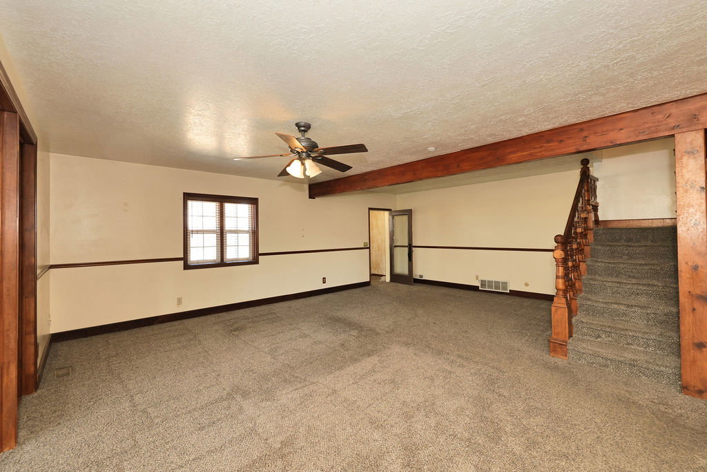 Spacious room featuring neutral carpeting, a ceiling fan, a window with grid panes, and exposed wood beam accents