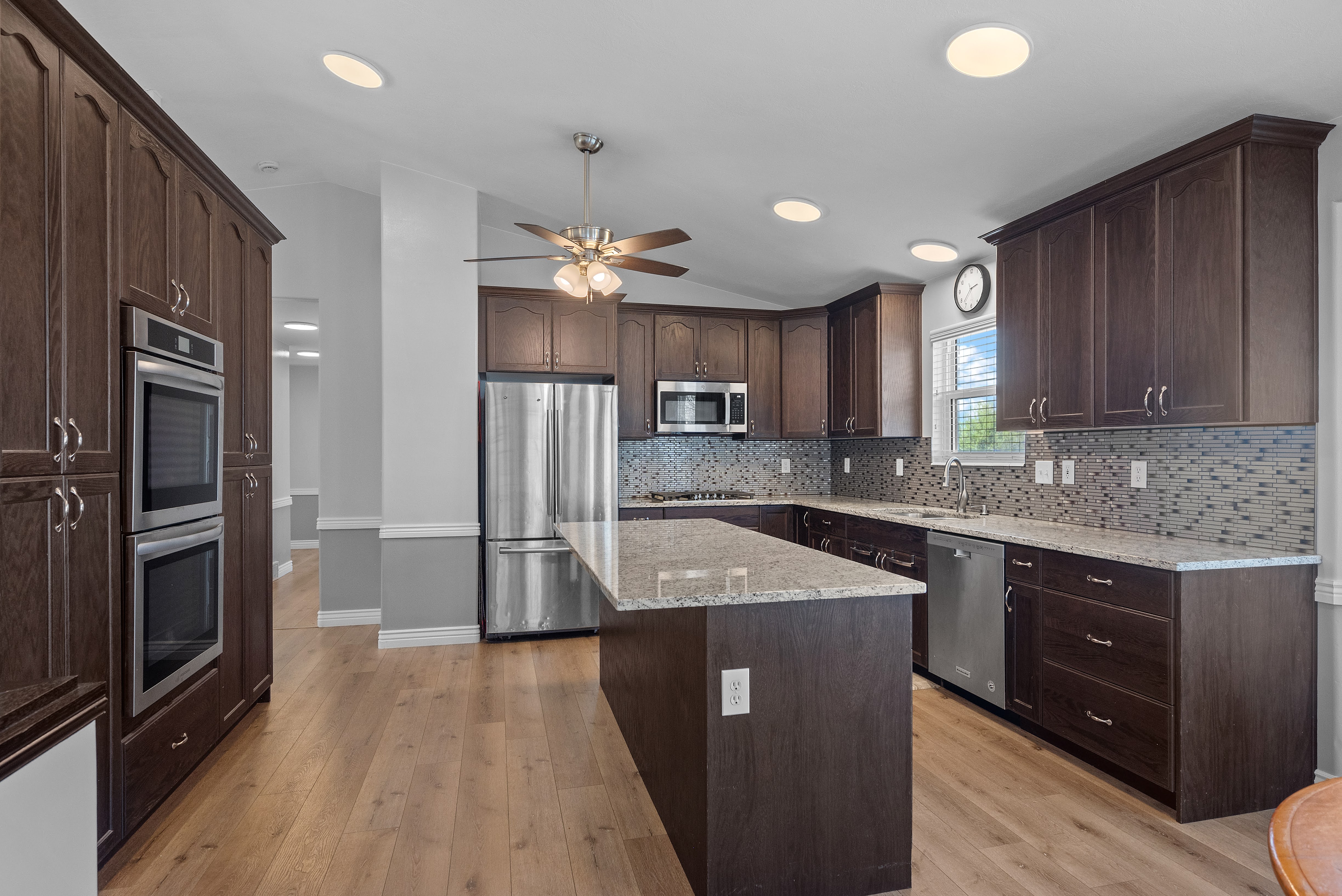 Kitchen featuring wood-finish flooring, dark wood cabinetry, stainless steel appliances, granite-style countertops, and a mosaic tile backsplash