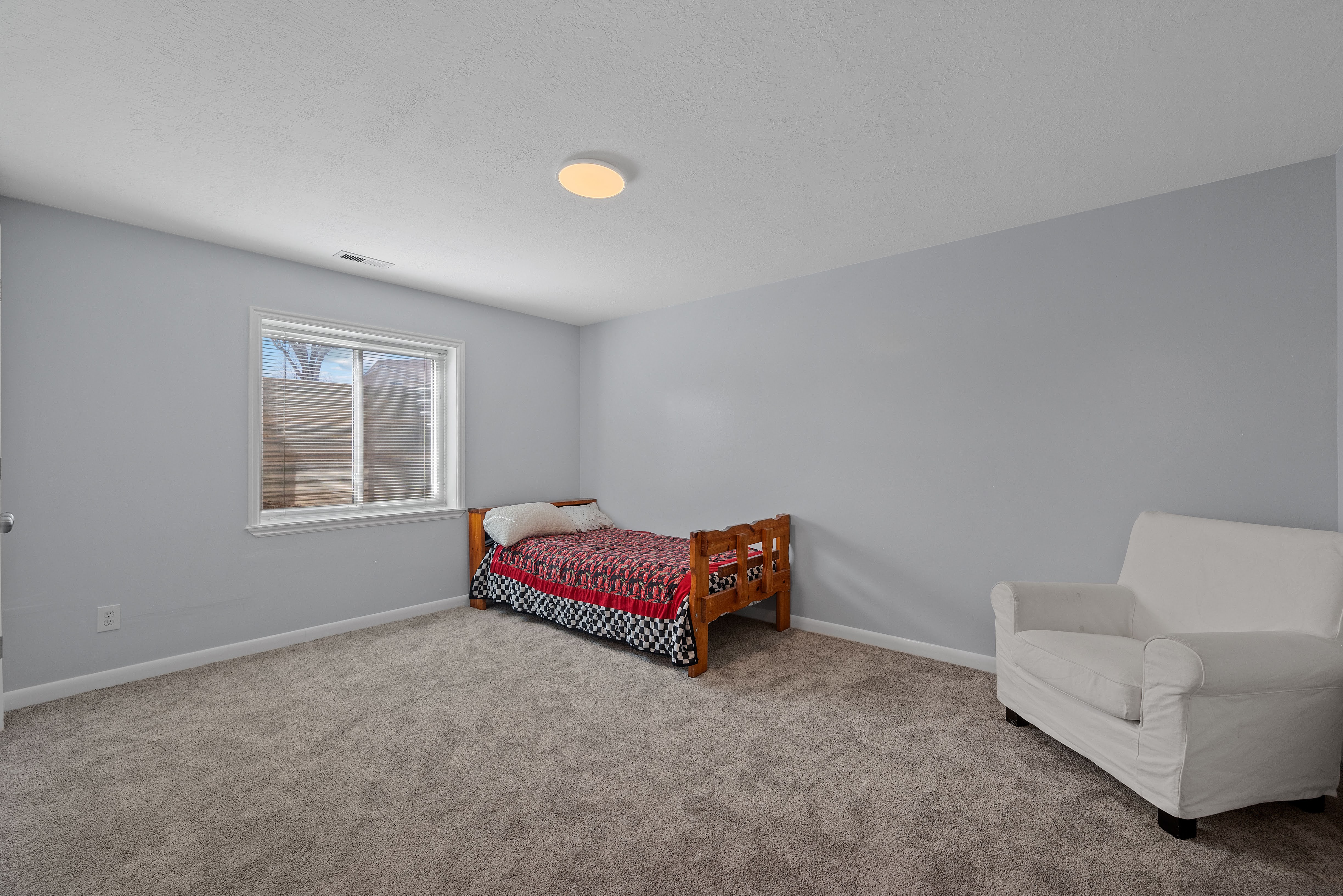 Spacious room featuring light gray walls, plush gray carpeting, a single window with blinds, and a ceiling-mounted light fixture
