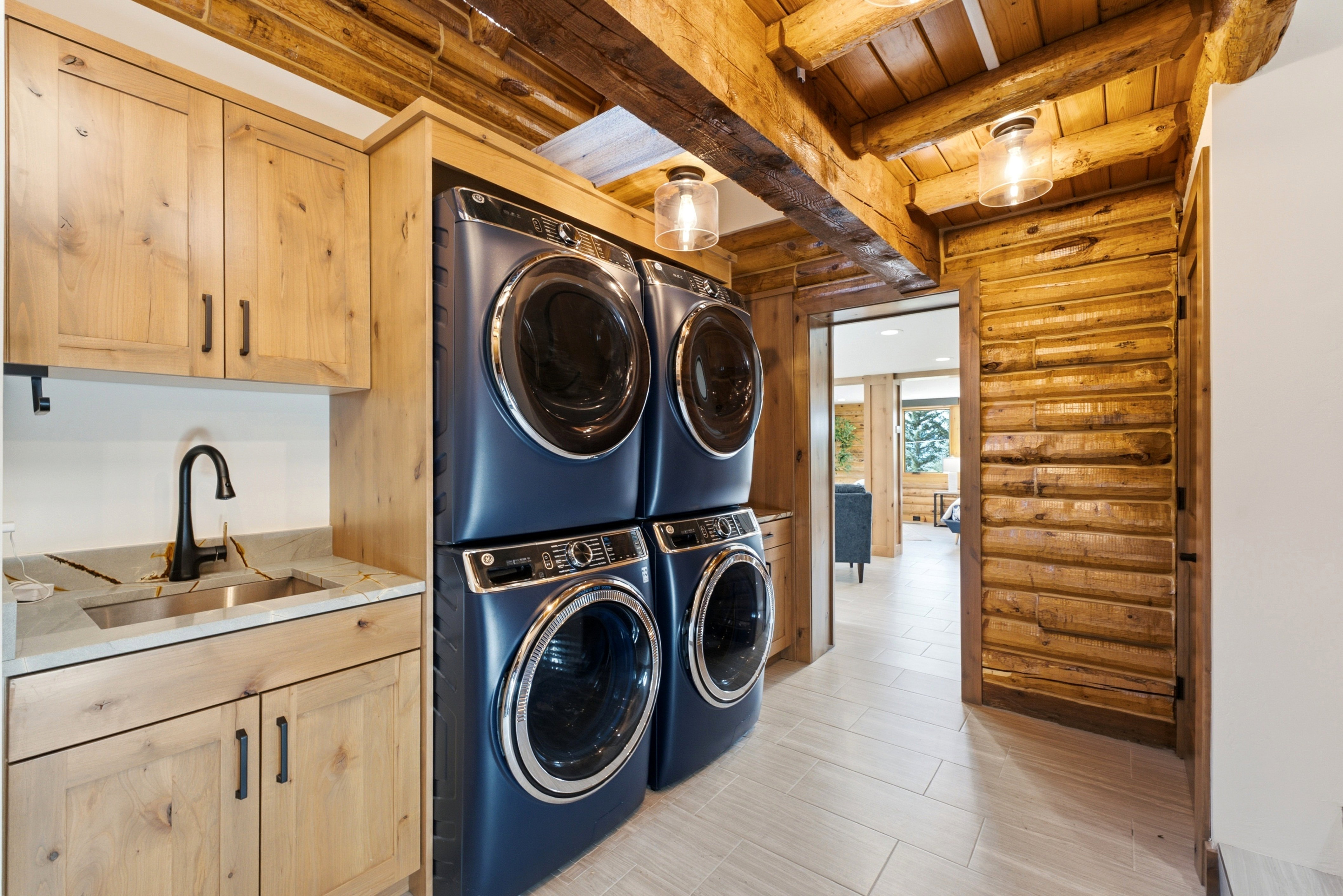 Laundry area featuring light wood cabinetry with dark hardware, a stone-finish countertop with an undermount sink and matte black faucet, and stacked front-load washers and dryers