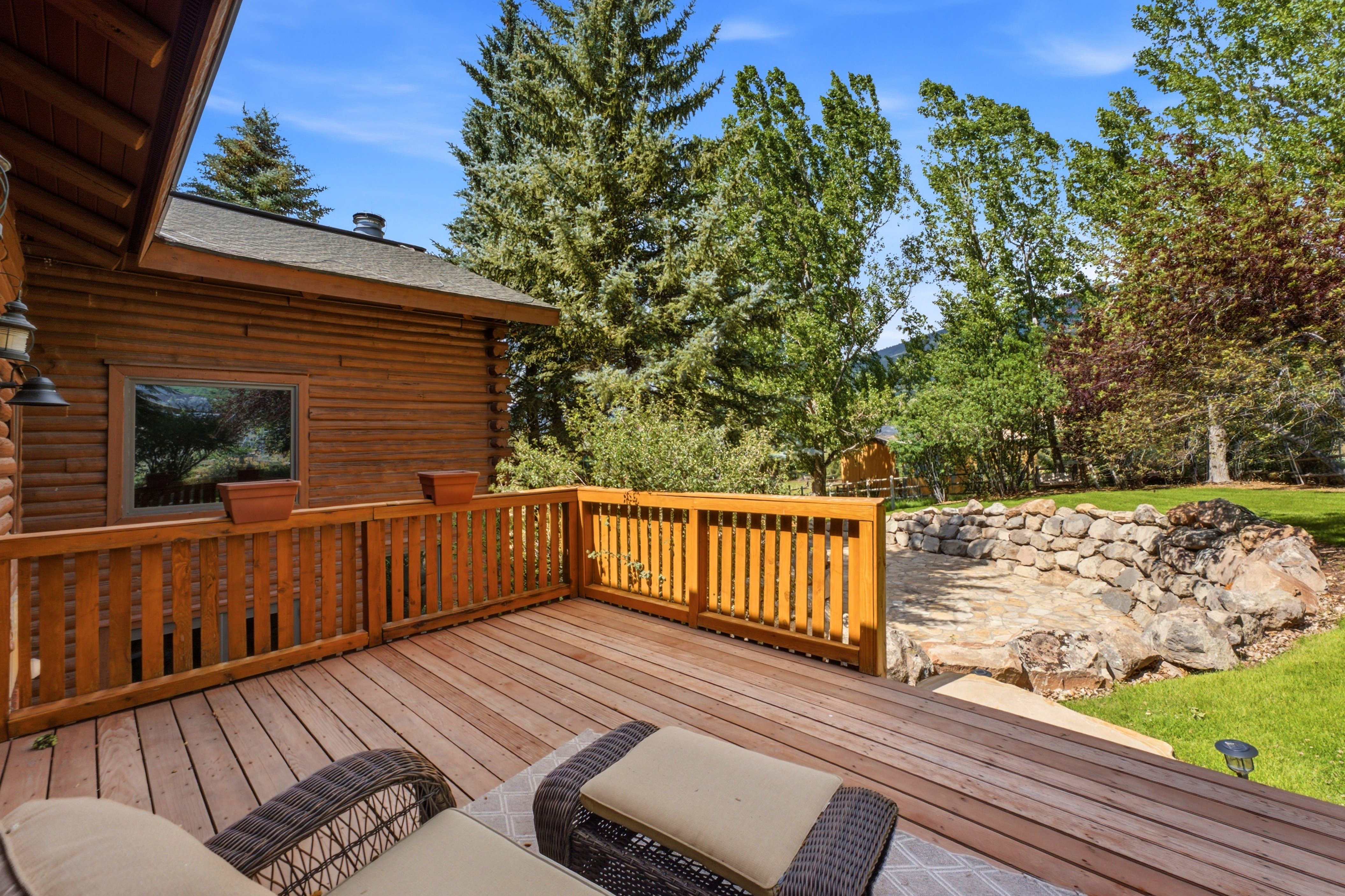 Expansive wood-finish deck with a wooden railing system, featuring an exterior log-finish wall, and a stone-lined patio area