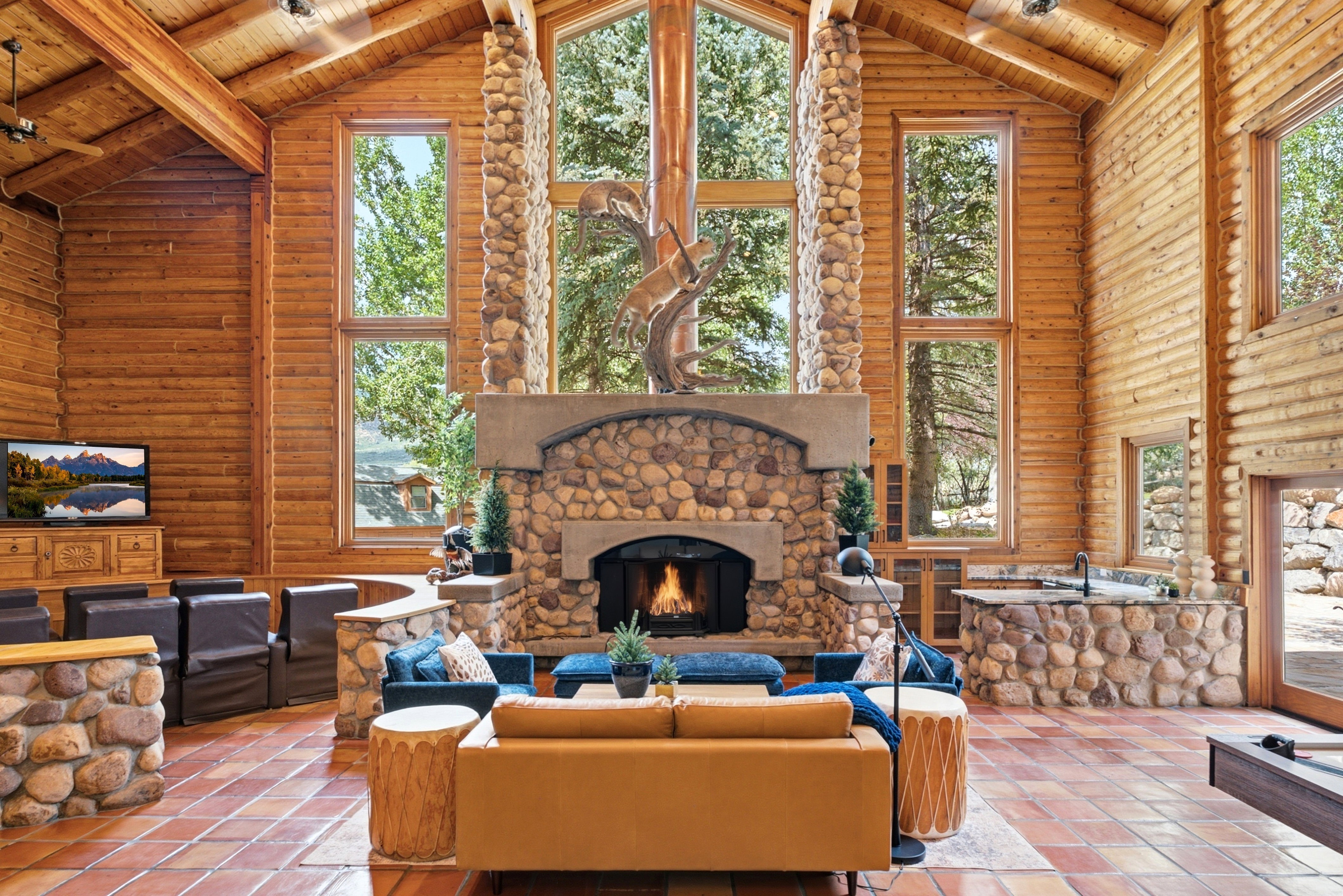 Vaulted ceiling with exposed wood beams, featuring a stacked stone fireplace, floor-to-ceiling windows, log wall construction, and terracotta-style floor tiling