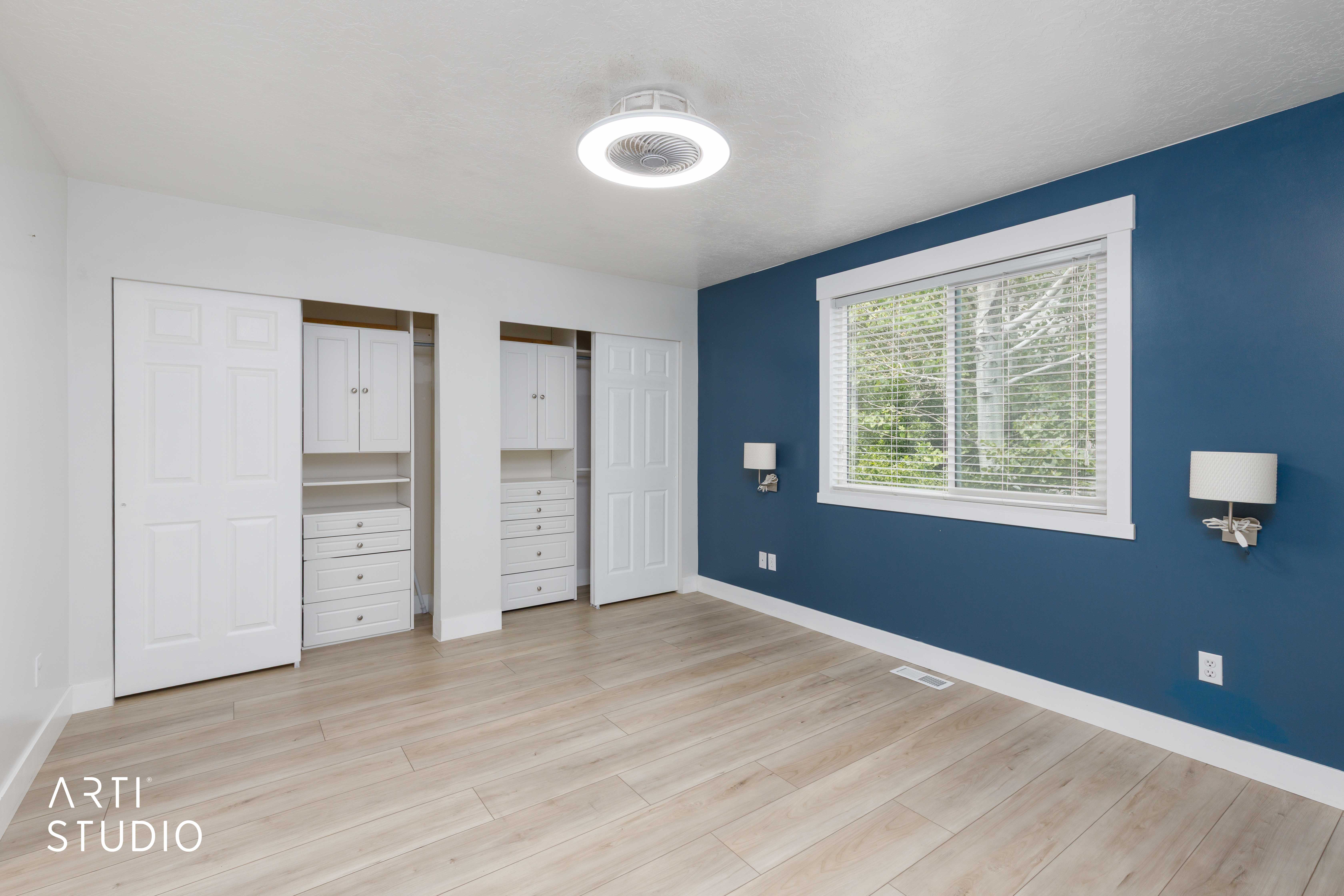Bedroom featuring light wood-finish flooring, a bright window with white blinds, and two built-in closet systems with drawers and cabinetry