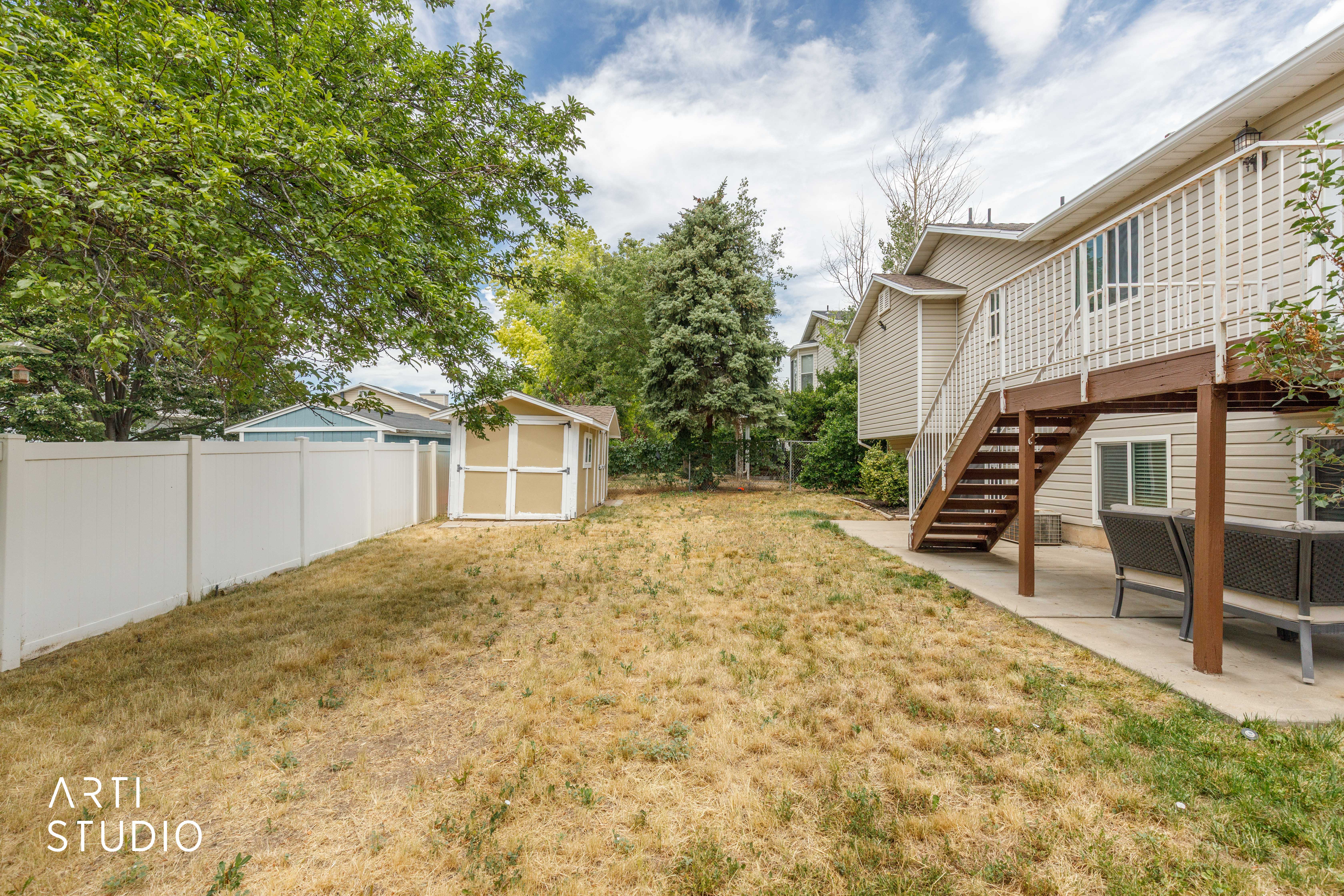 Spacious backyard featuring a built-in storage shed, a white privacy fence, mature trees, and a raised wooden deck with stairs providing outdoor access
