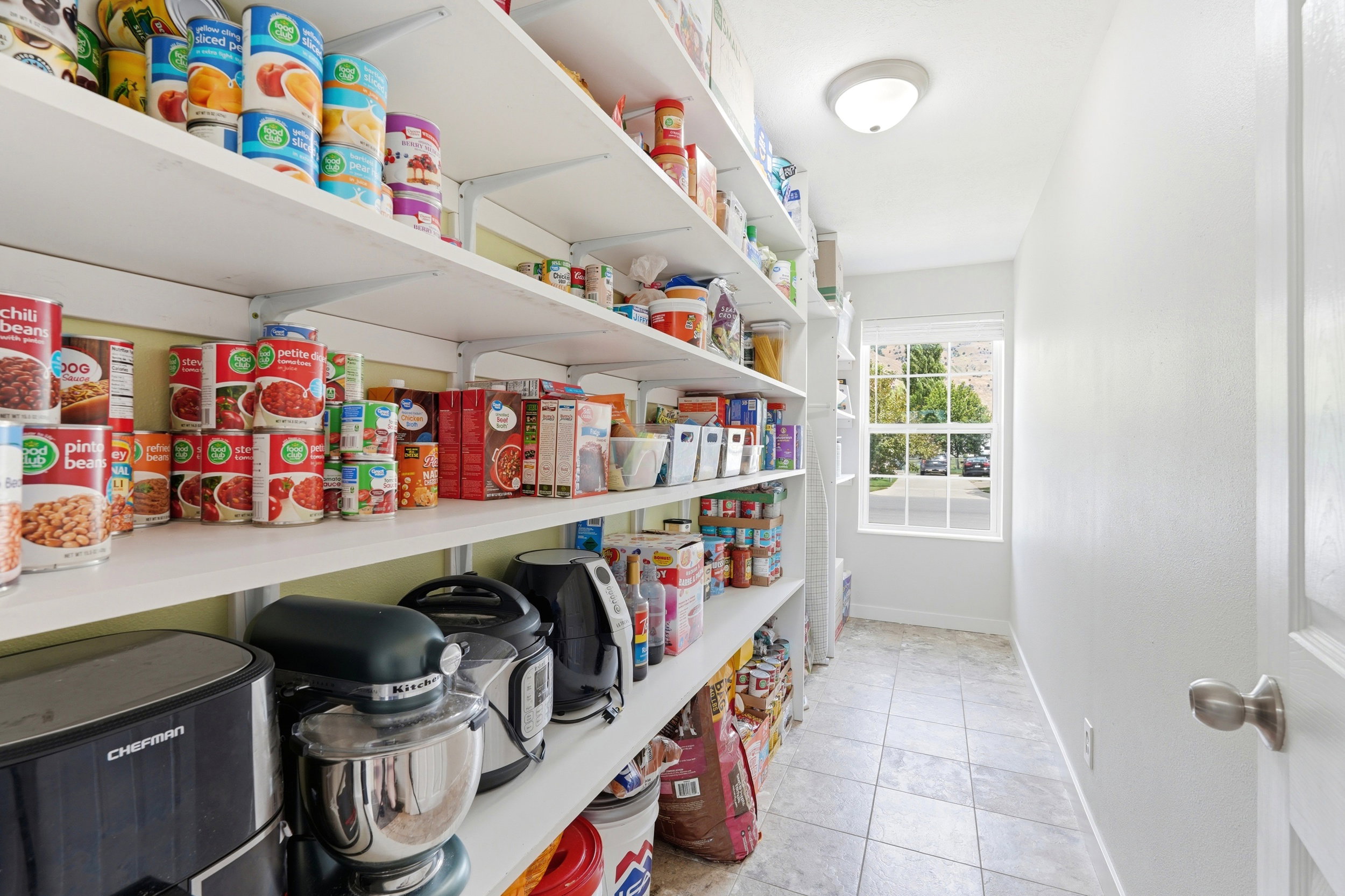 Spacious pantry with extensive shelving, square paned window, overhead flush-mount lighting, and neutral tile flooring