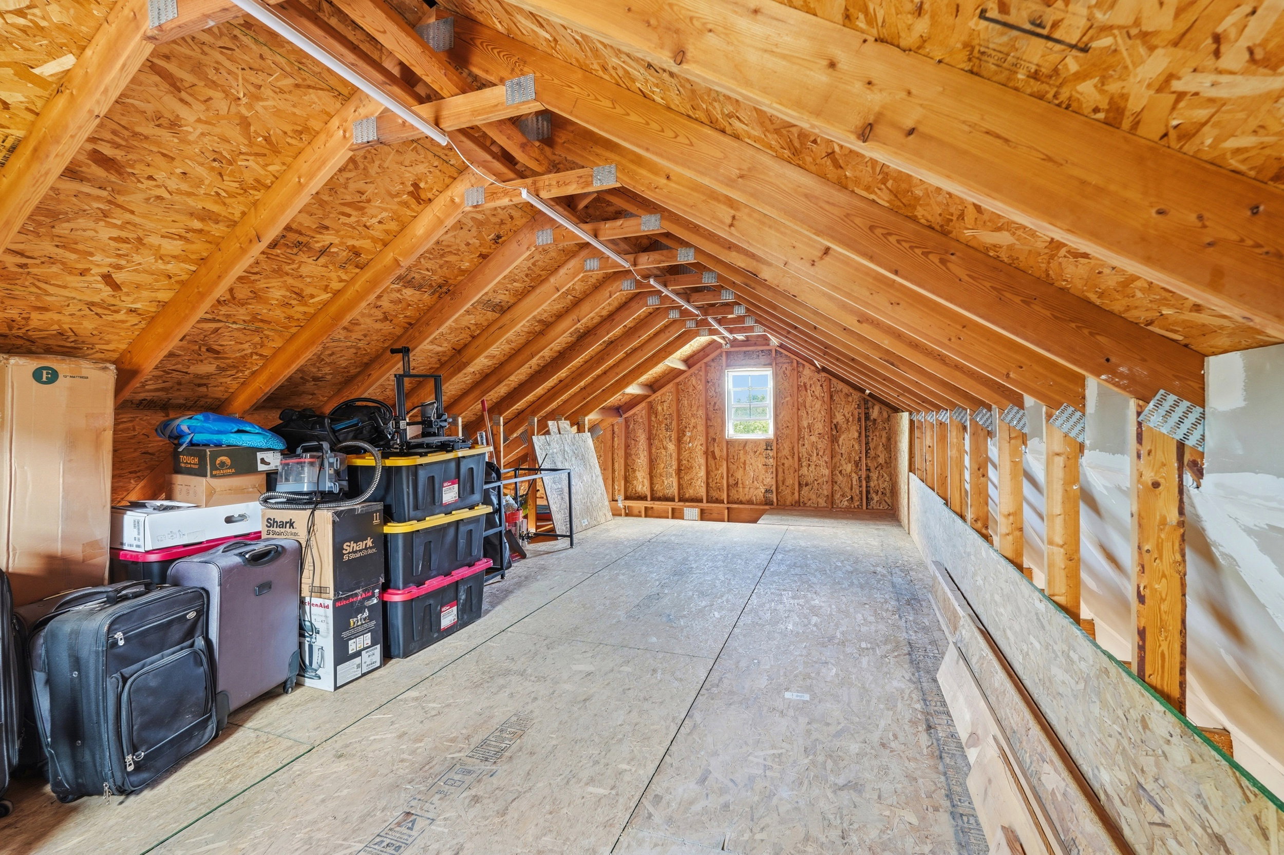 Spacious attic featuring exposed wood rafters, plywood flooring, a single window, and a fluorescent light fixture