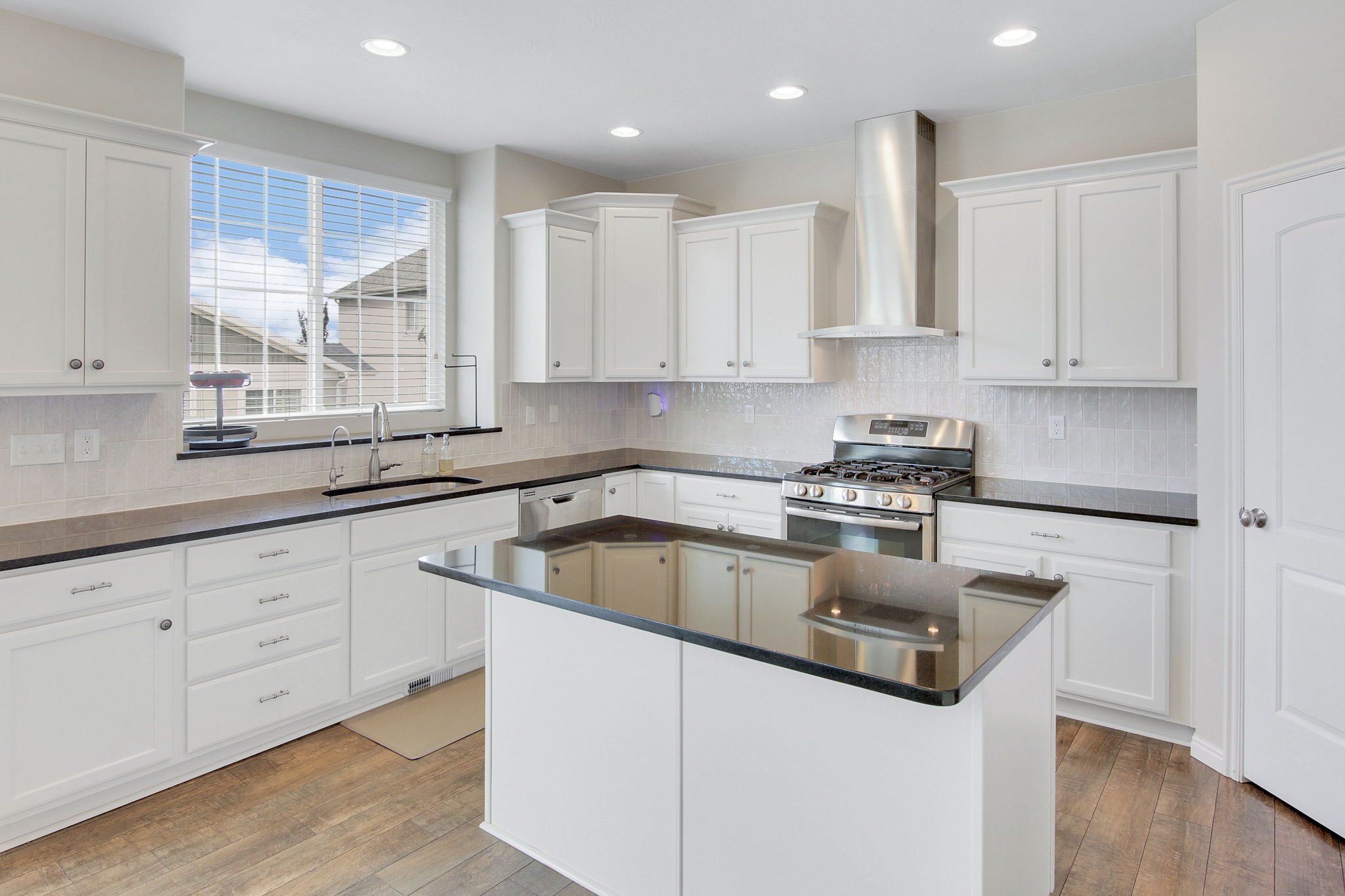 Contemporary kitchen featuring an island with a black countertop, white shaker-style cabinetry, stainless steel range with matching vent hood, recessed lighting, and wood-finish flooring