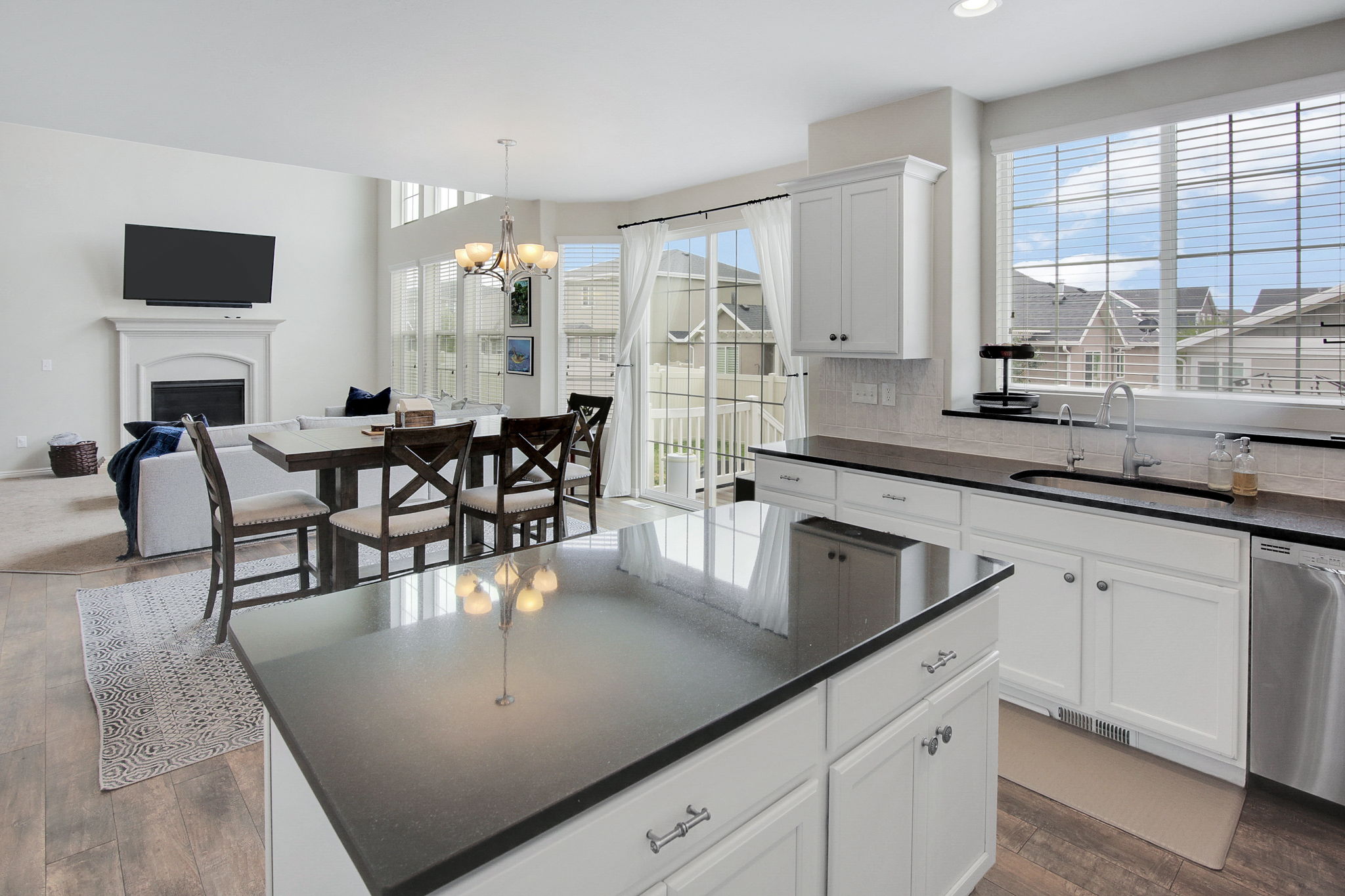 Open-concept living space featuring wood-finish flooring, a kitchen island with dark countertops, white cabinetry, a large window over the sink, and a fireplace with a white mantel