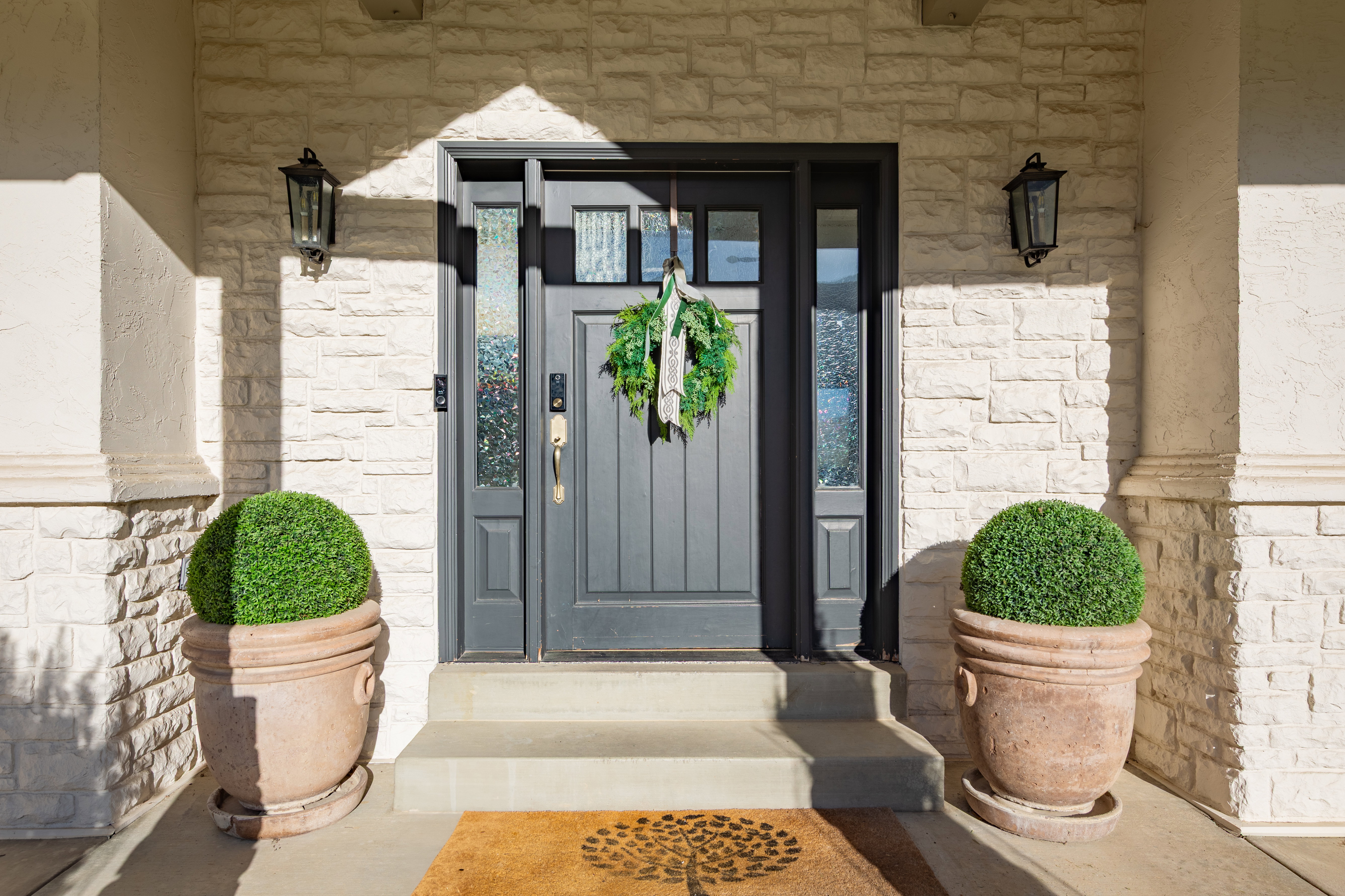 Entryway featuring a dark gray paneled door with glass sidelights, textured stone veneer, stucco columns, concrete steps, and two black wall-mounted lanterns