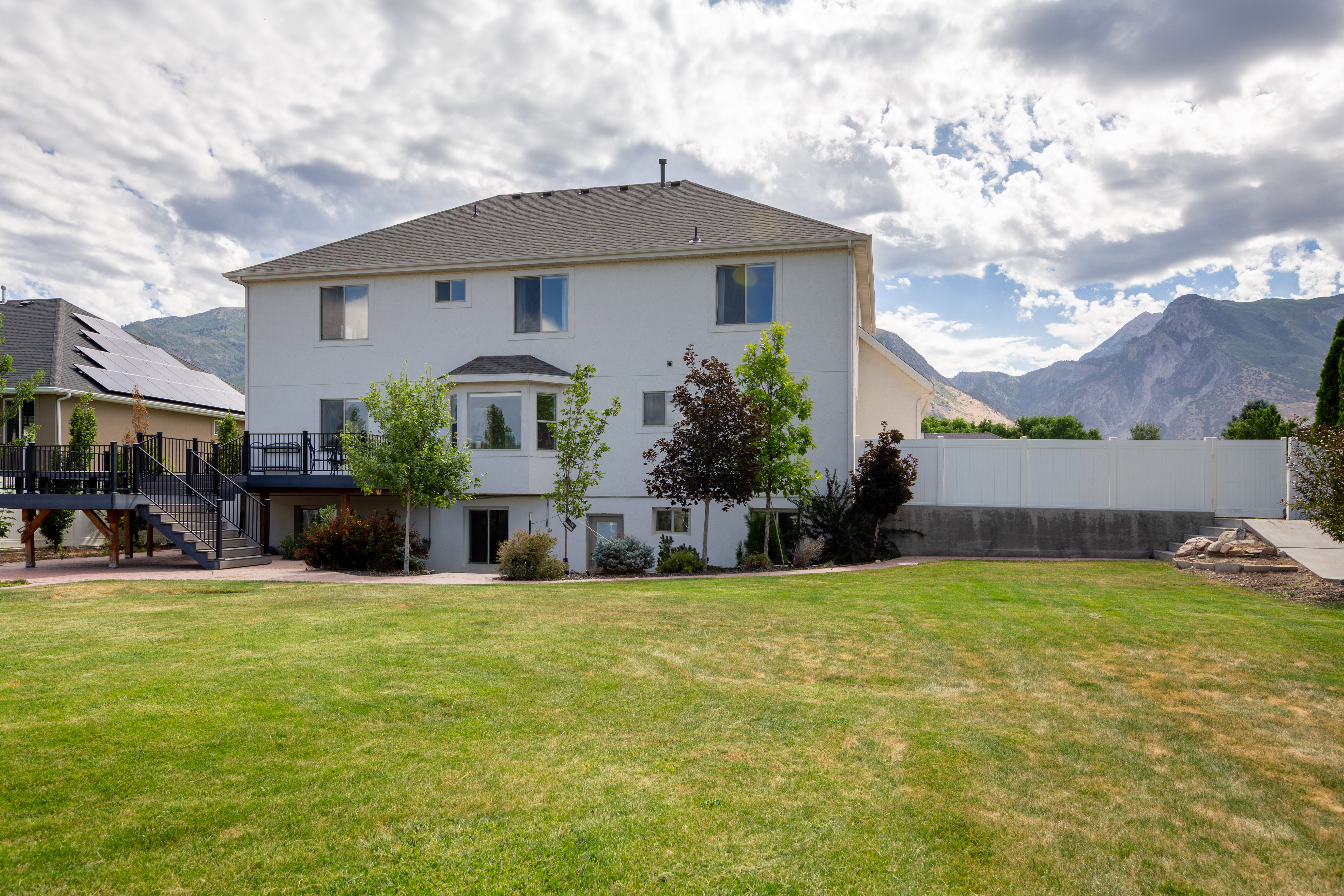 Expansive backyard featuring a manicured lawn, a two-story residence with a bay window, a raised deck with stairs, a white privacy fence, and mountain scenery