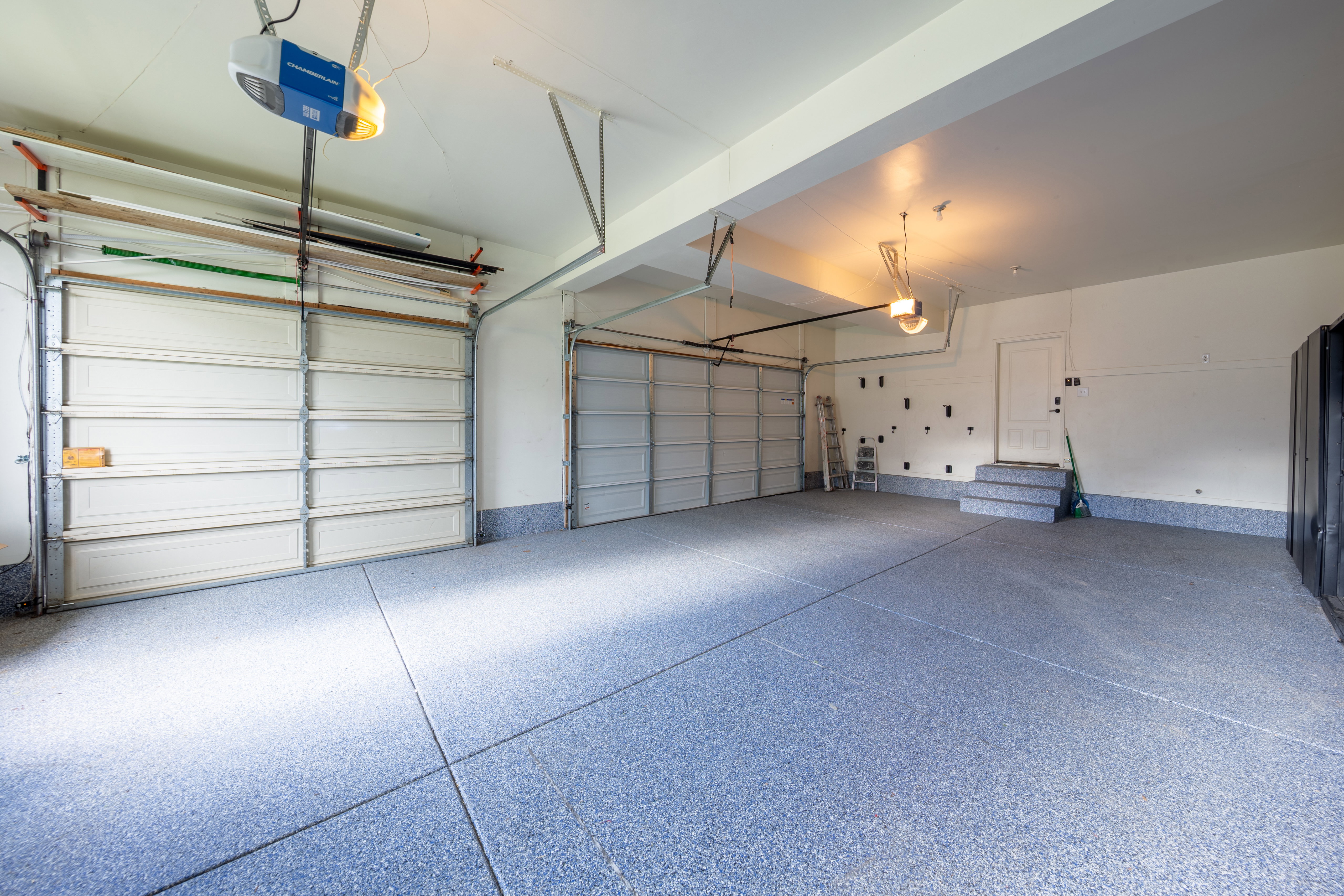 Spacious garage featuring a durable speckled epoxy floor, two overhead garage doors, overhead garage door openers, and a utility door with a two-step entry