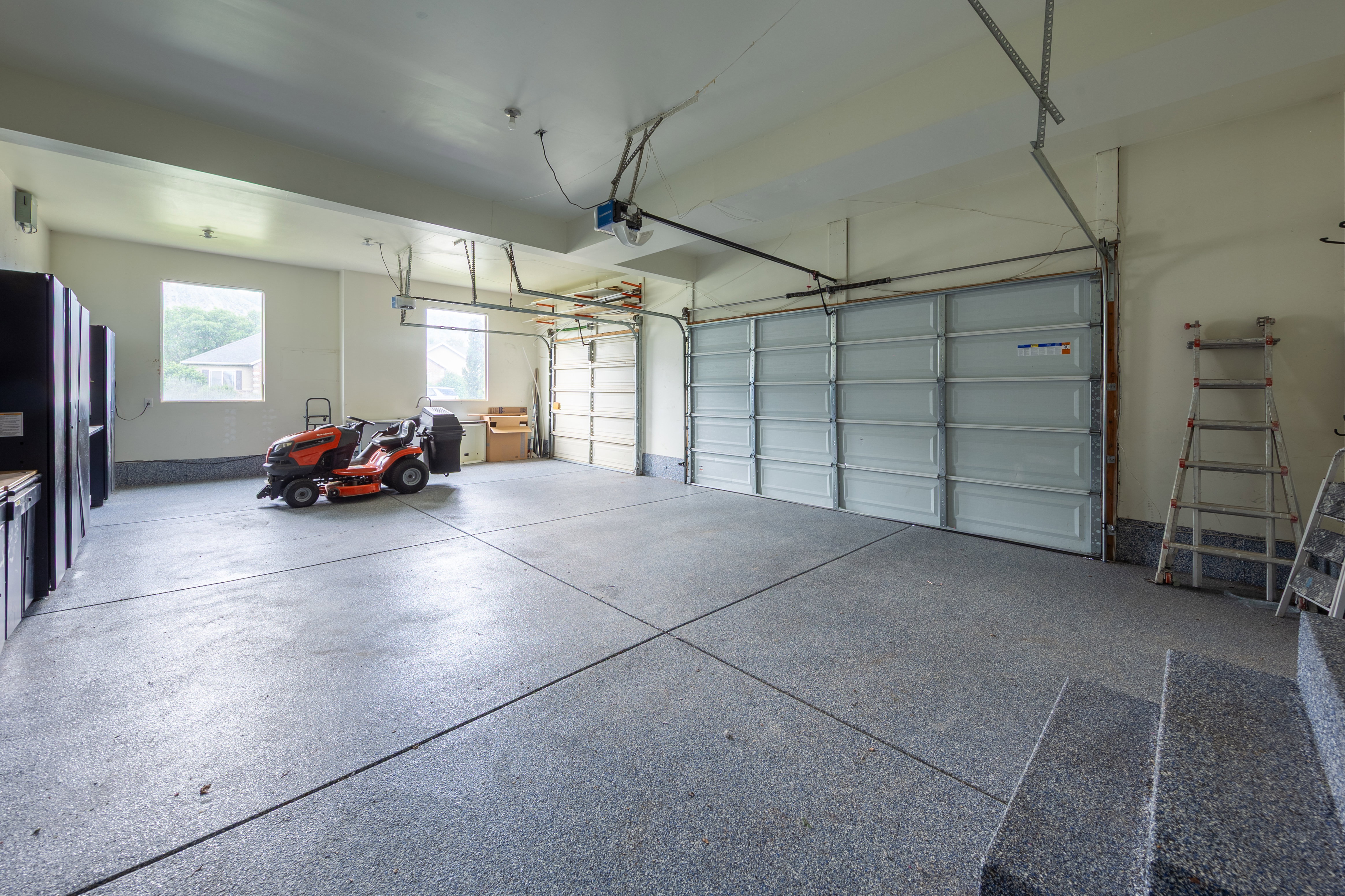 Spacious garage featuring a durable epoxy-coated floor, dual overhead garage doors, built-in cabinetry with a wood-finish countertop, and multiple windows providing natural light