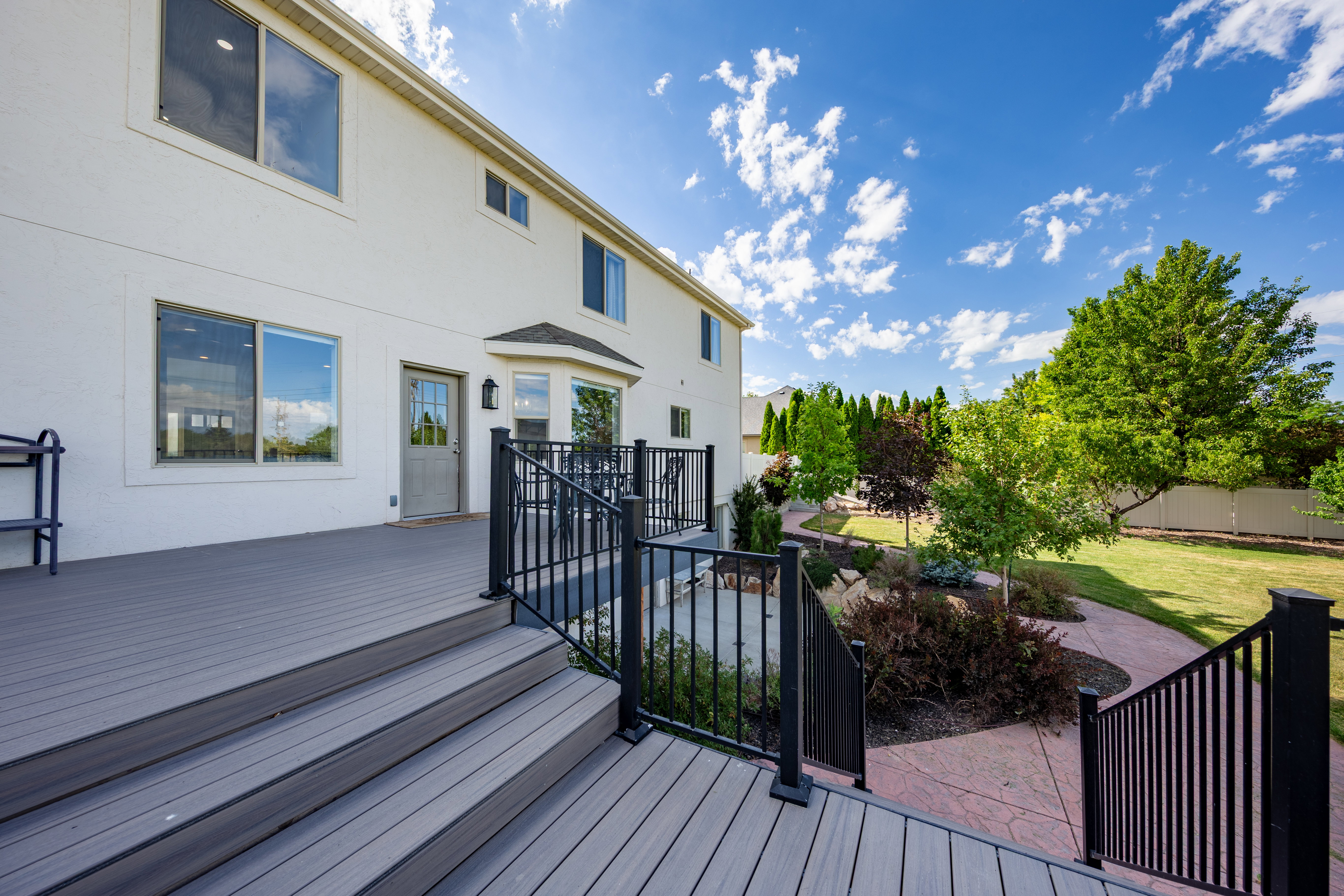 Expansive composite deck with black metal railings, offering direct access to a landscaped backyard featuring mature trees, a manicured lawn, and a stamped concrete pathway