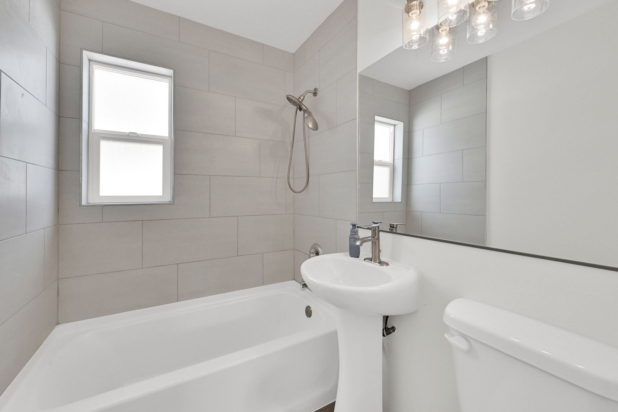 Contemporary bathroom featuring light gray subway tile surrounding the tub, a white pedestal sink with brushed nickel faucet, a frameless wall mirror, and modern vanity lighting
