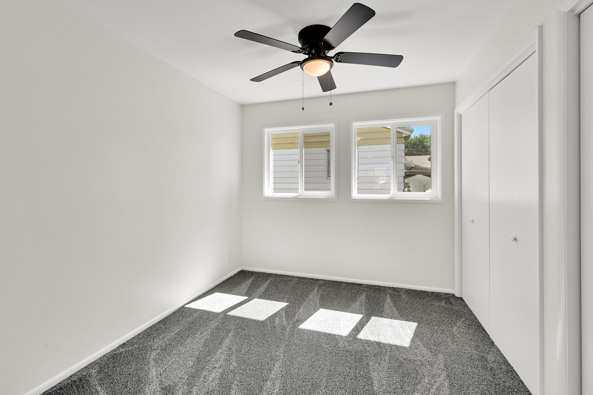 Room featuring a ceiling fan with integrated lighting, two windows with white frames, dark gray carpet flooring, and white bi-fold closet doors