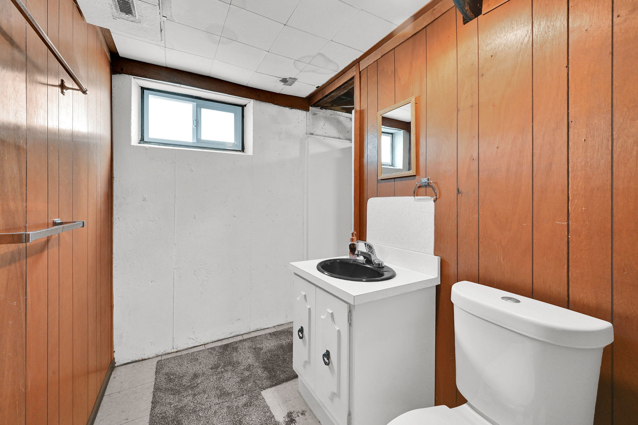 Compact bathroom featuring wood-panel walls, a white vanity with a black sink, a toilet, and a small window