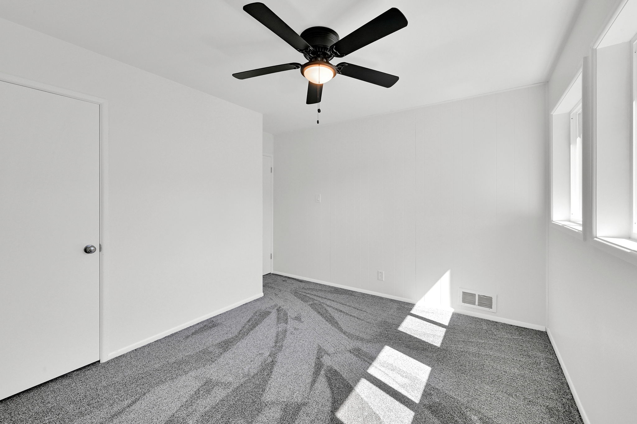 Bright room featuring a ceiling fan with integrated lighting, two windows, wall-to-wall carpeting, and a white interior door