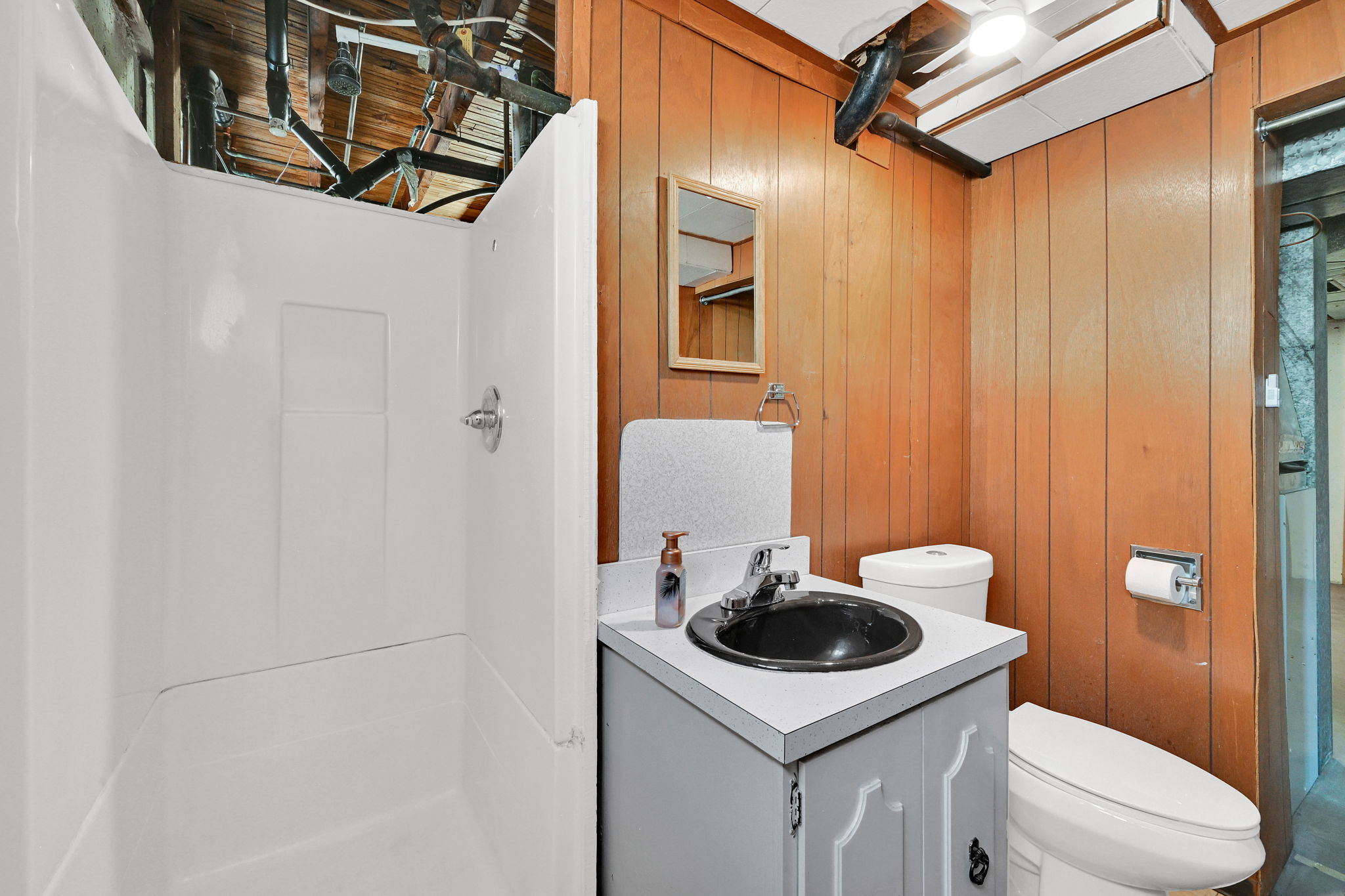 Compact bathroom featuring a white shower stall with chrome fixtures, a gray vanity with a black basin sink, and wood-paneled walls