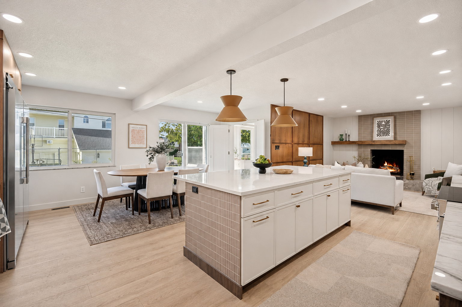 Open-concept living space featuring a central kitchen island with white cabinetry, wood-finish flooring, recessed lighting, and a prominent fireplace with a light brick surround and wood mantel