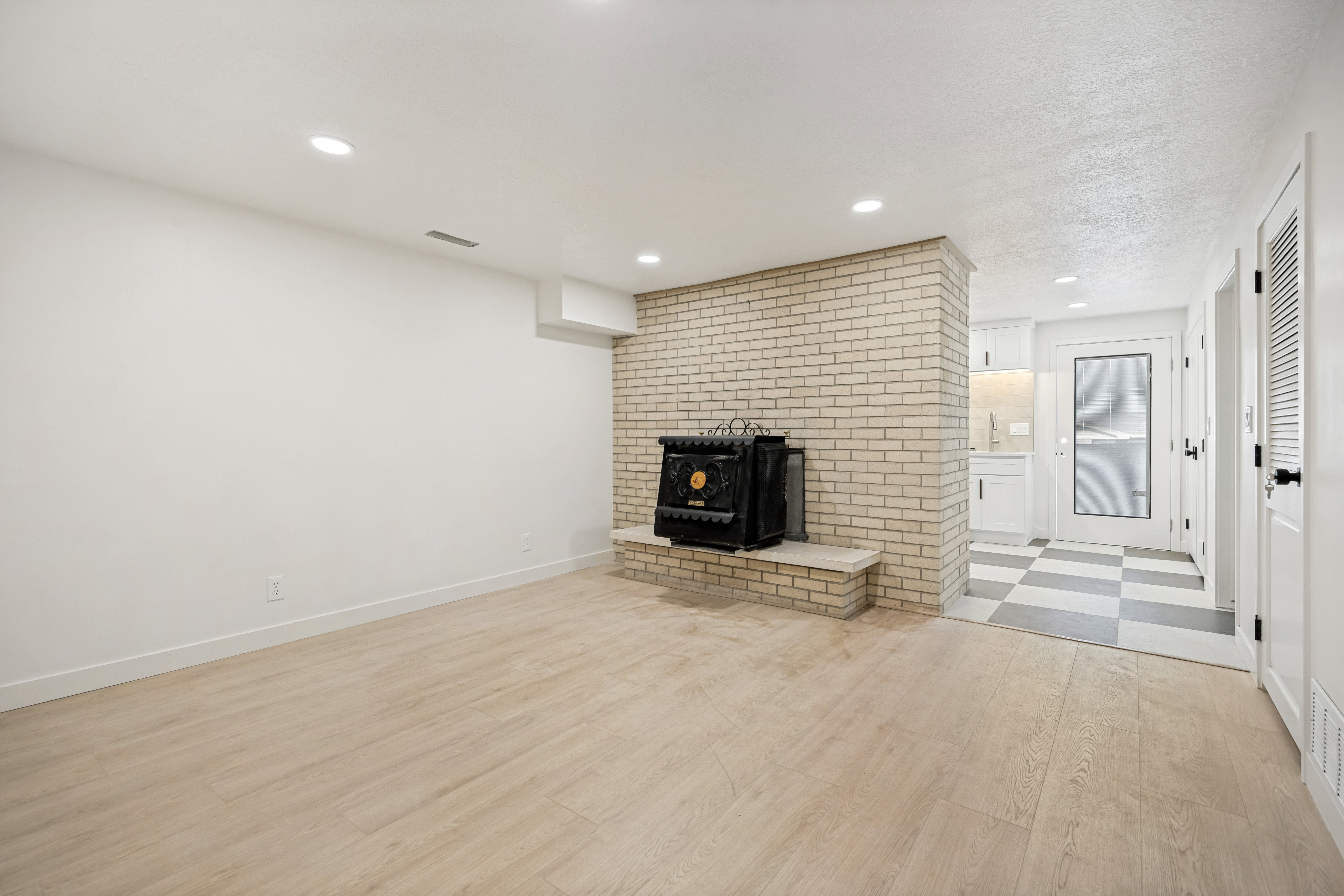Spacious room featuring light wood-finish flooring, recessed lighting, and a prominent brick-clad fireplace with a black stove insert