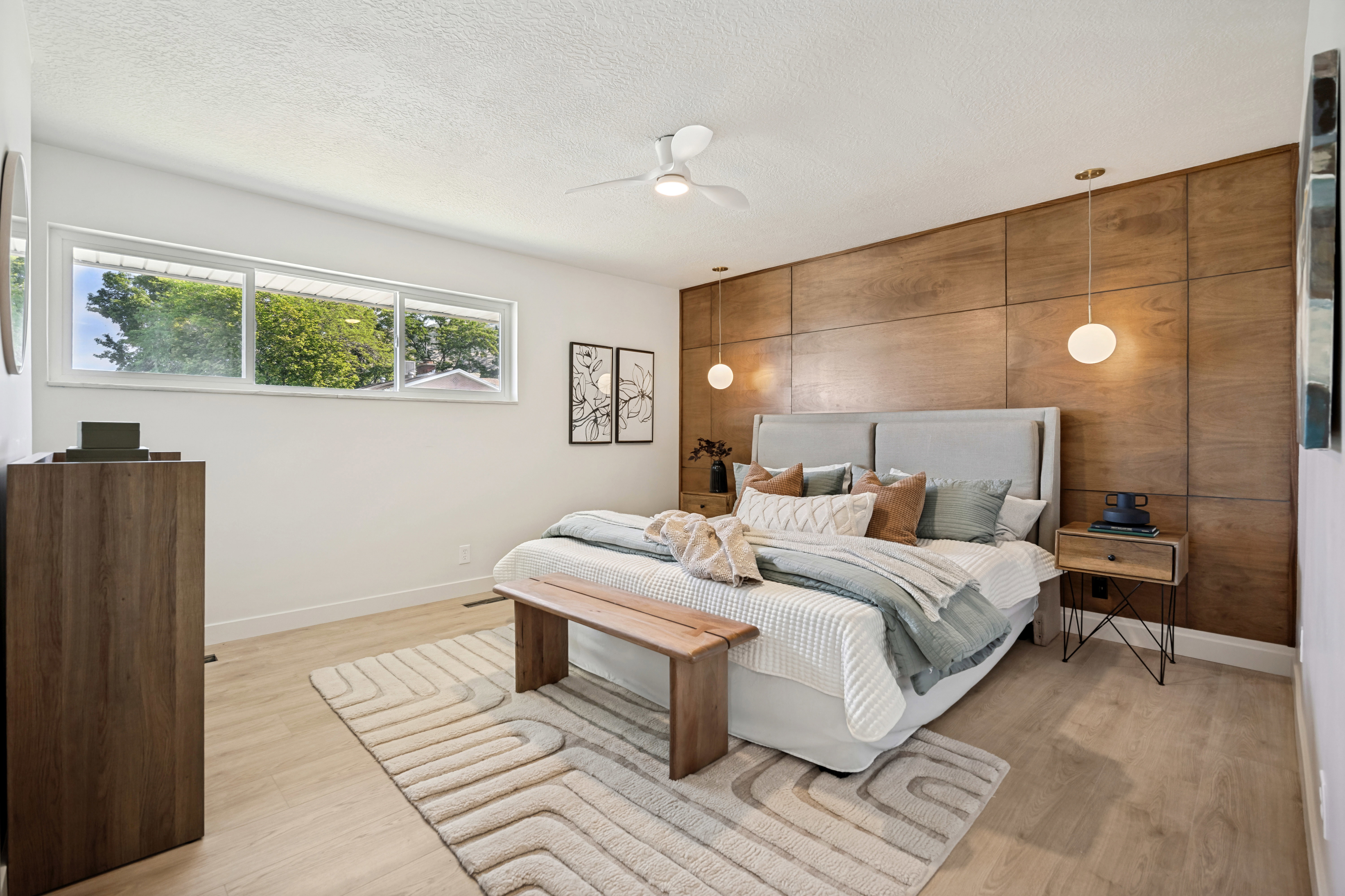 Bedroom featuring a full wood-paneled accent wall, light wood-finish flooring, a ceiling fan with integrated lighting, and a wide horizontal window