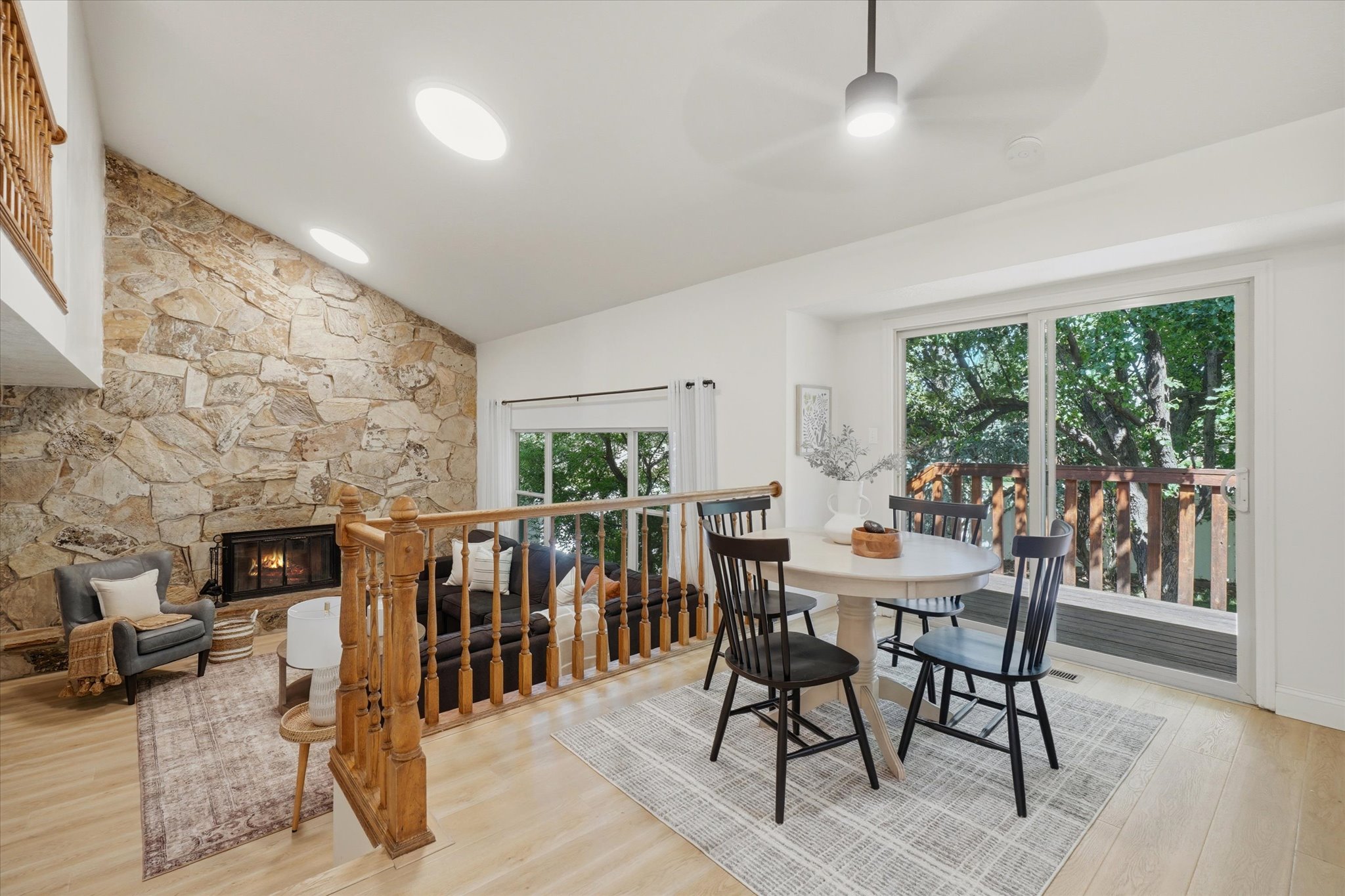Vaulted ceiling with skylights, stone fireplace, light wood-finish flooring, and sliding glass door leading to an outdoor balcony