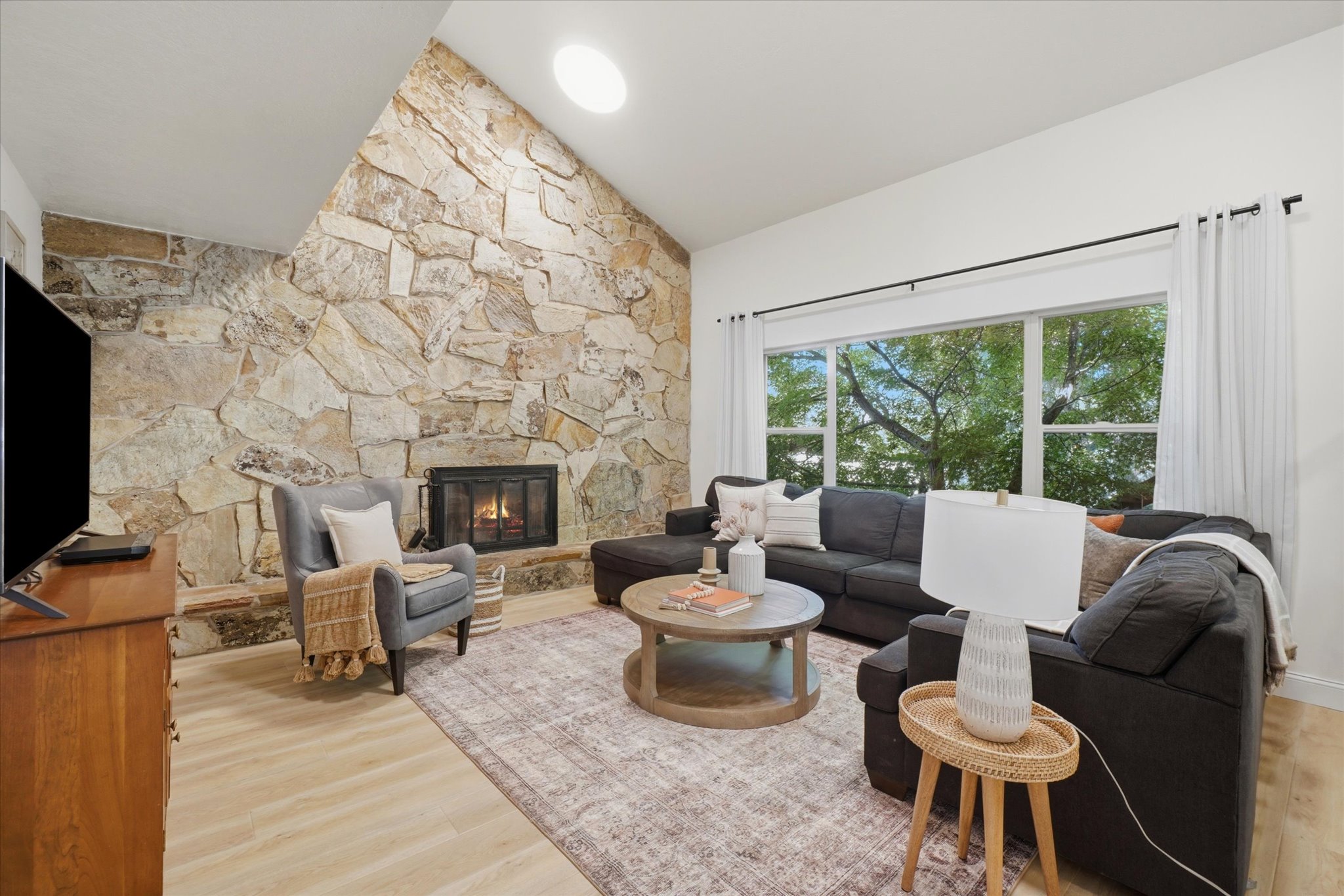 Vaulted ceiling living area featuring a full-height stone accent wall, an integrated fireplace, expansive window, a circular skylight, and light wood-finish flooring