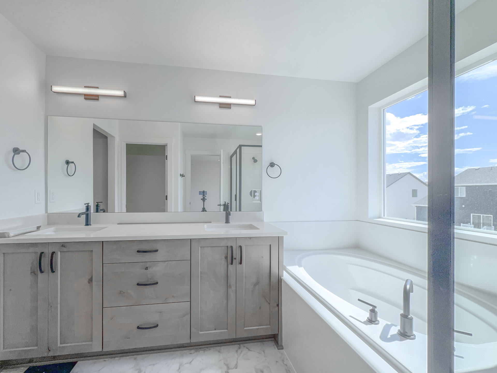 Spacious bathroom featuring a dual vanity with light wood-finish cabinetry, built-in soaking tub, large window, and matte black fixtures