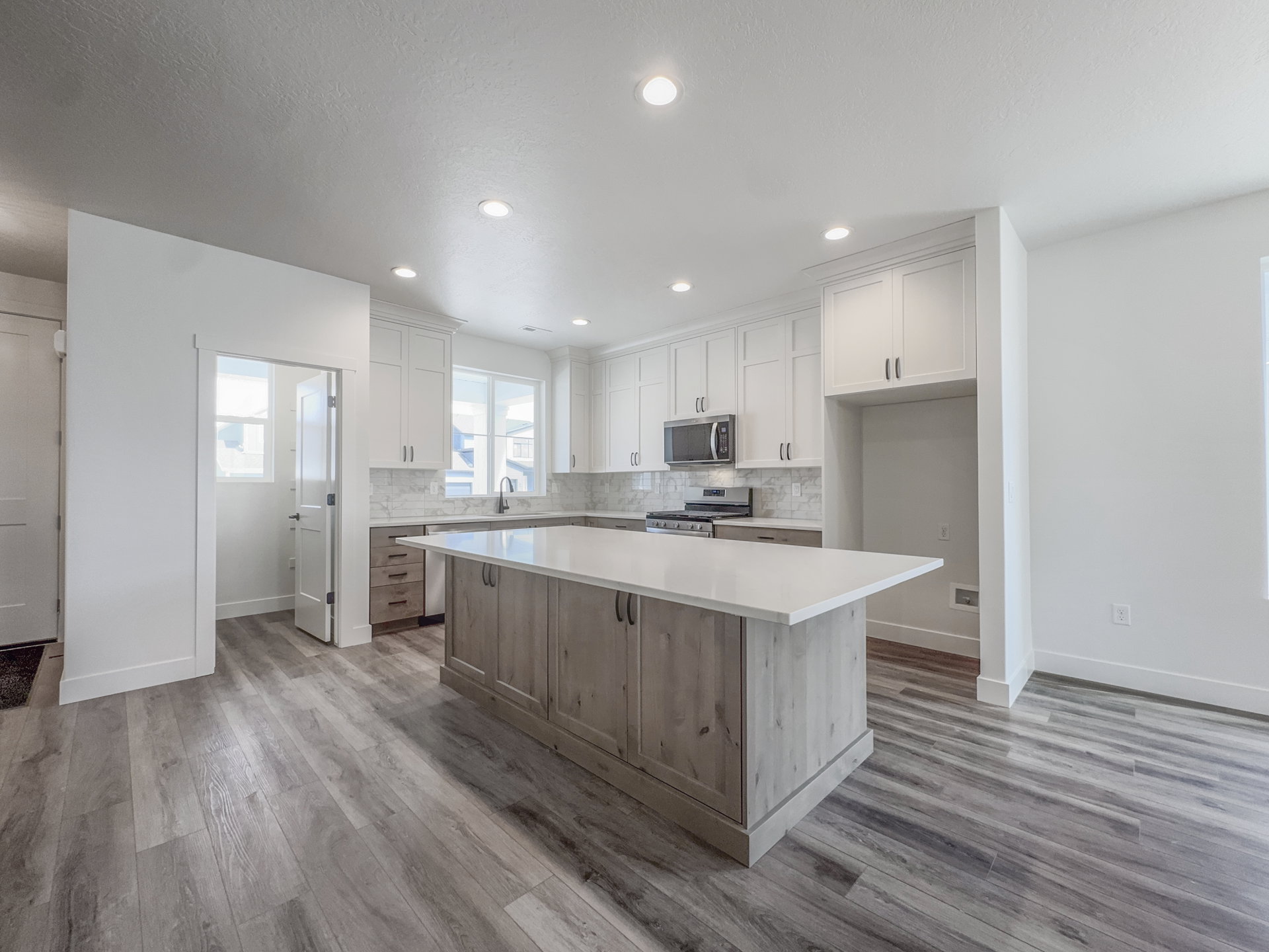 Contemporary kitchen featuring a large central island with a light-toned countertop, white upper cabinetry, grey lower cabinetry, stainless steel appliances, and wood-finish flooring