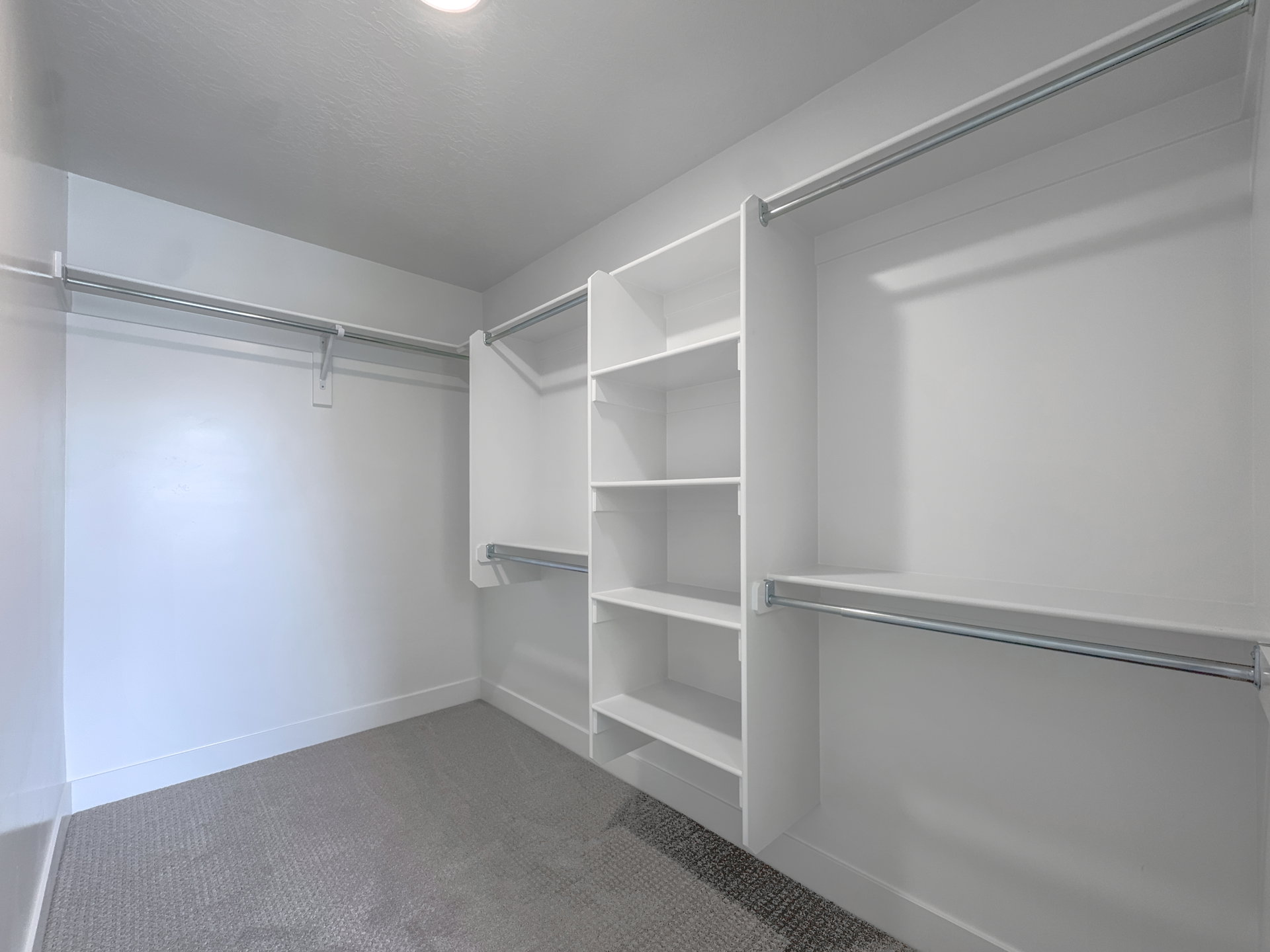 Spacious walk-in closet featuring white wall finishes, built-in shelving, chrome hanging rods, recessed ceiling lighting, and gray carpet flooring