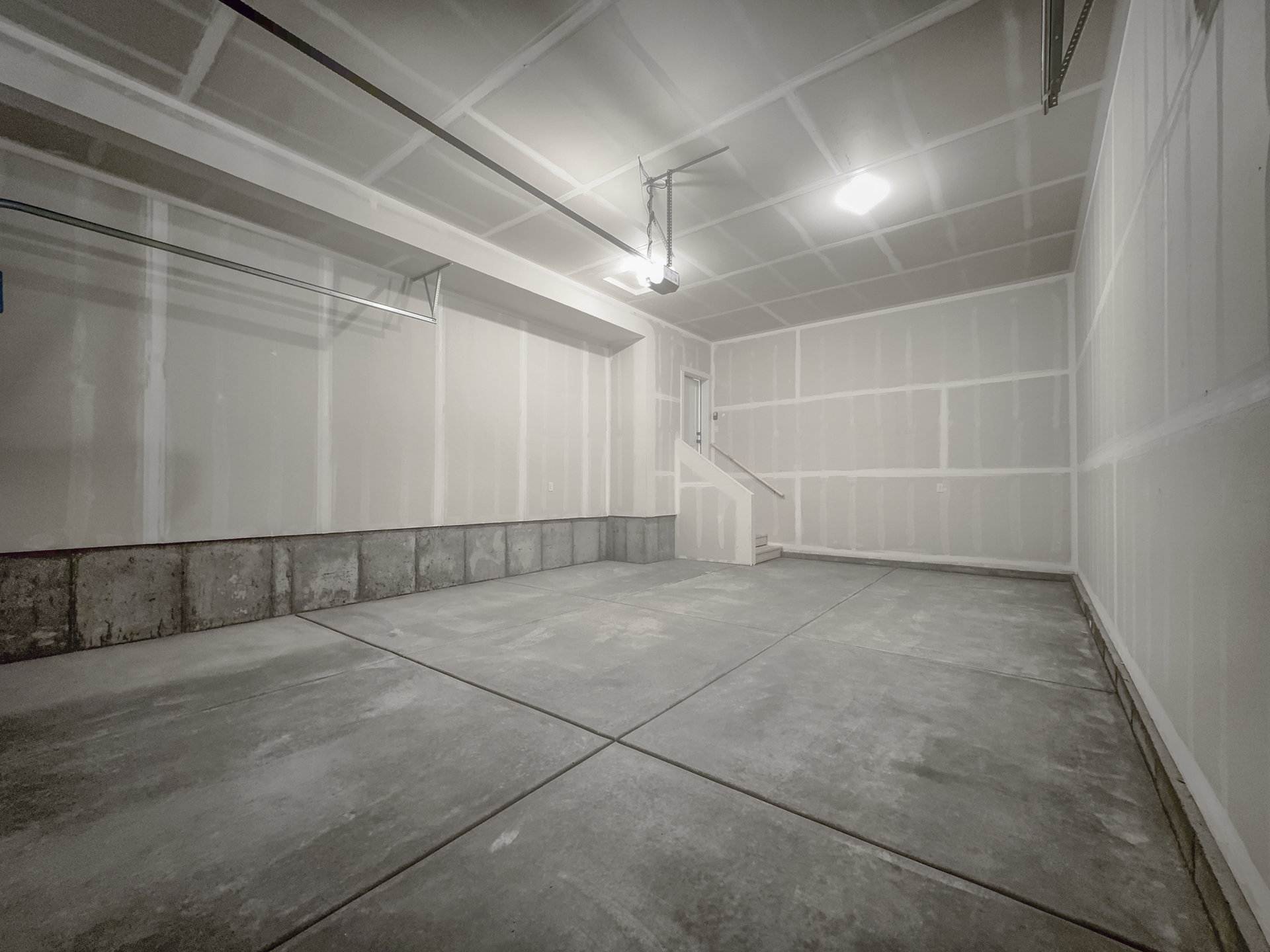 Spacious garage featuring a concrete floor, exposed drywall, overhead lighting, and an access door with stairs