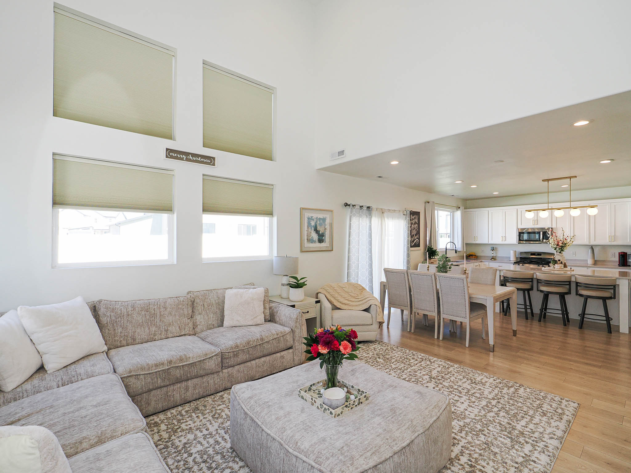 Vaulted ceiling living area featuring high windows with blinds, wood-finish flooring, and an open-concept kitchen with white cabinetry, stainless steel appliances, and a central island with bar seating