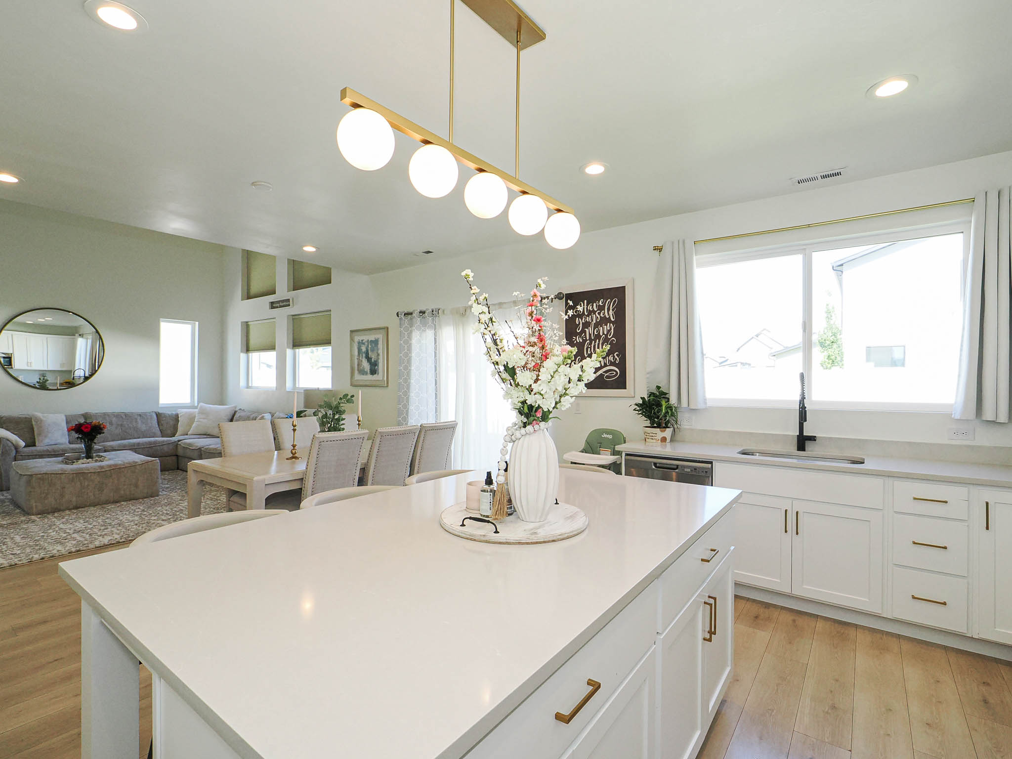 Open-concept living space featuring a central kitchen island with a light-toned countertop, white cabinetry with gold-finish hardware, wood-finish flooring, and a dining area