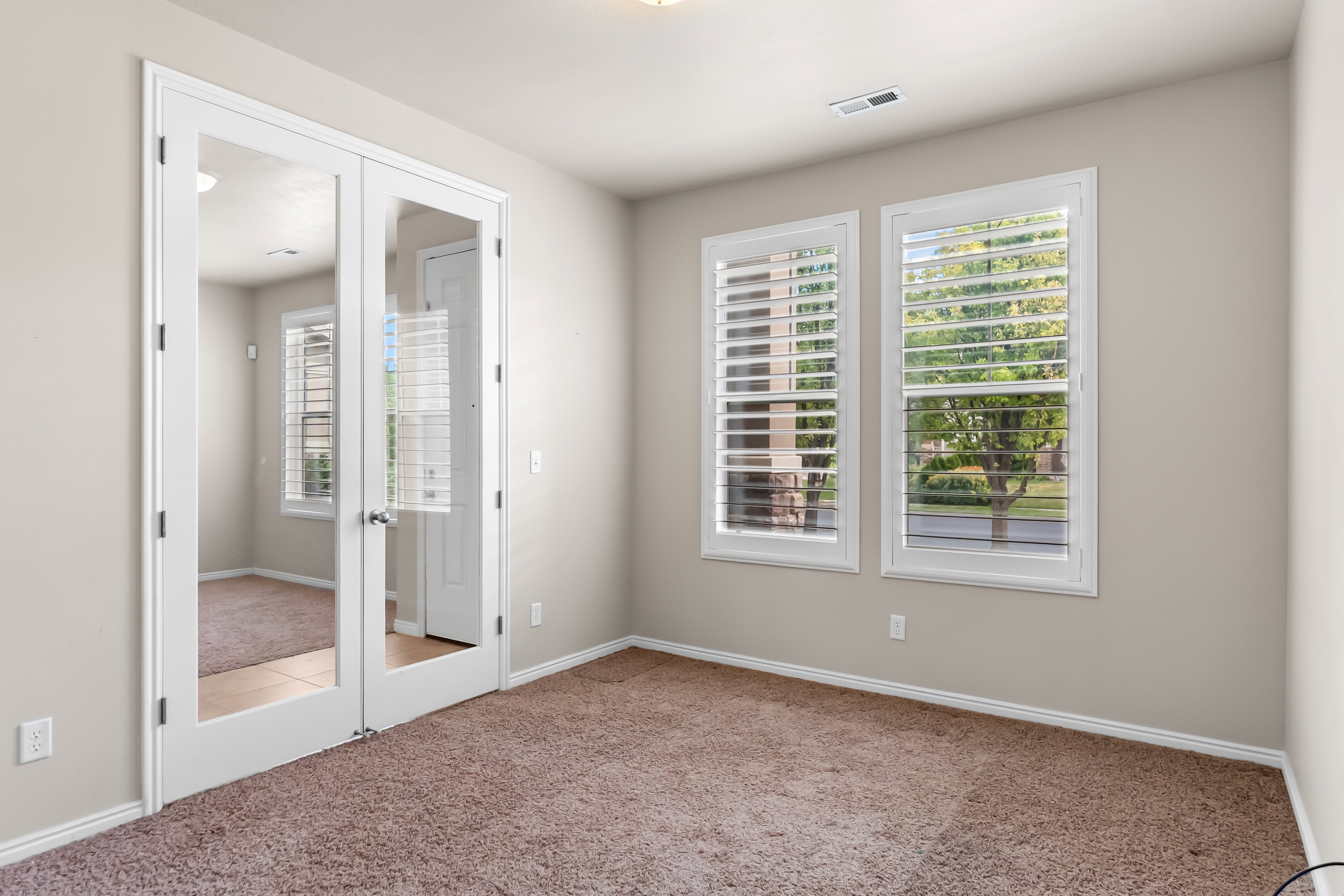 Room with white French doors, two windows featuring white louvered shutters, light beige wall paint, and brown carpet flooring