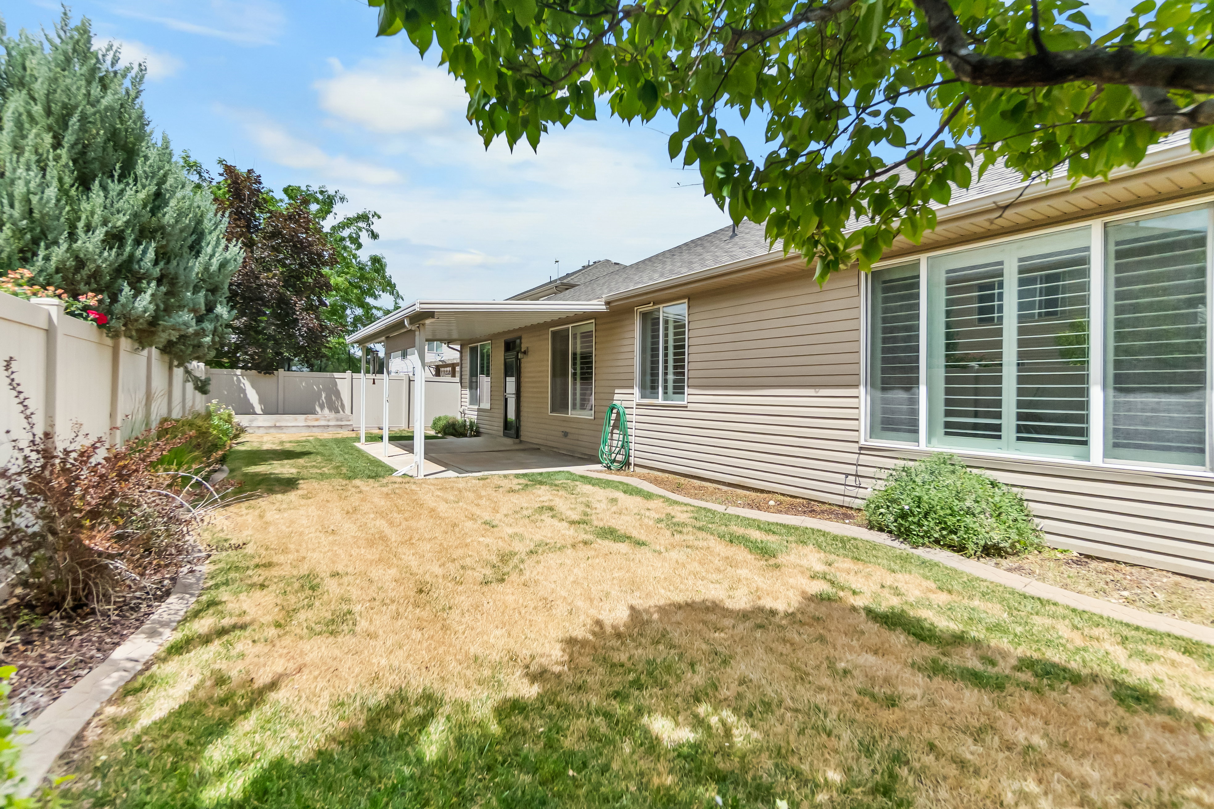 Expansive backyard featuring a covered patio, concrete hardscaping, vinyl fencing, mature landscaping, and bay windows with plantation shutters