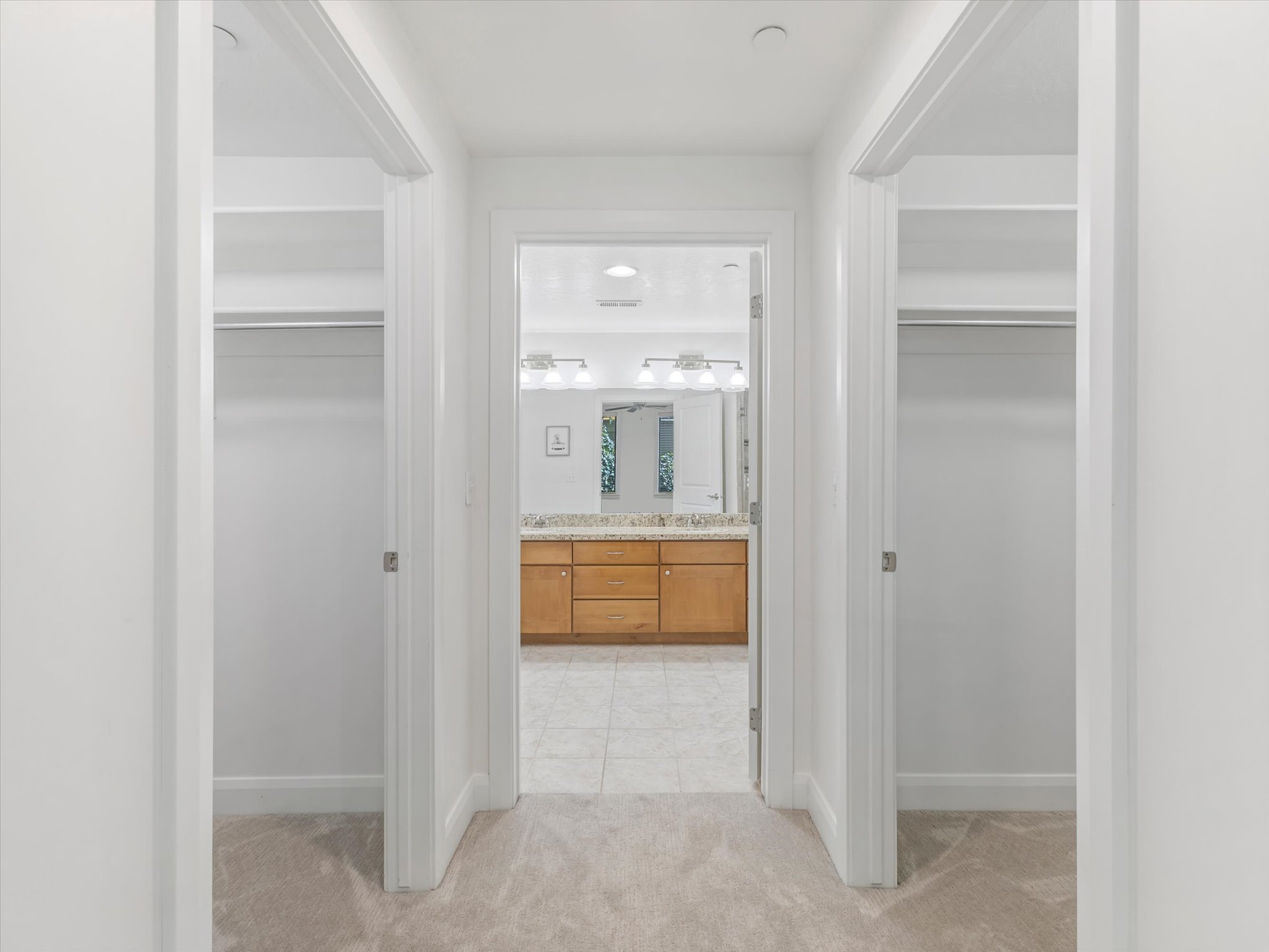 Hallway with light-toned carpet flooring, leading to a bathroom featuring a dual vanity with a light wood-finish cabinet and a light-toned countertop