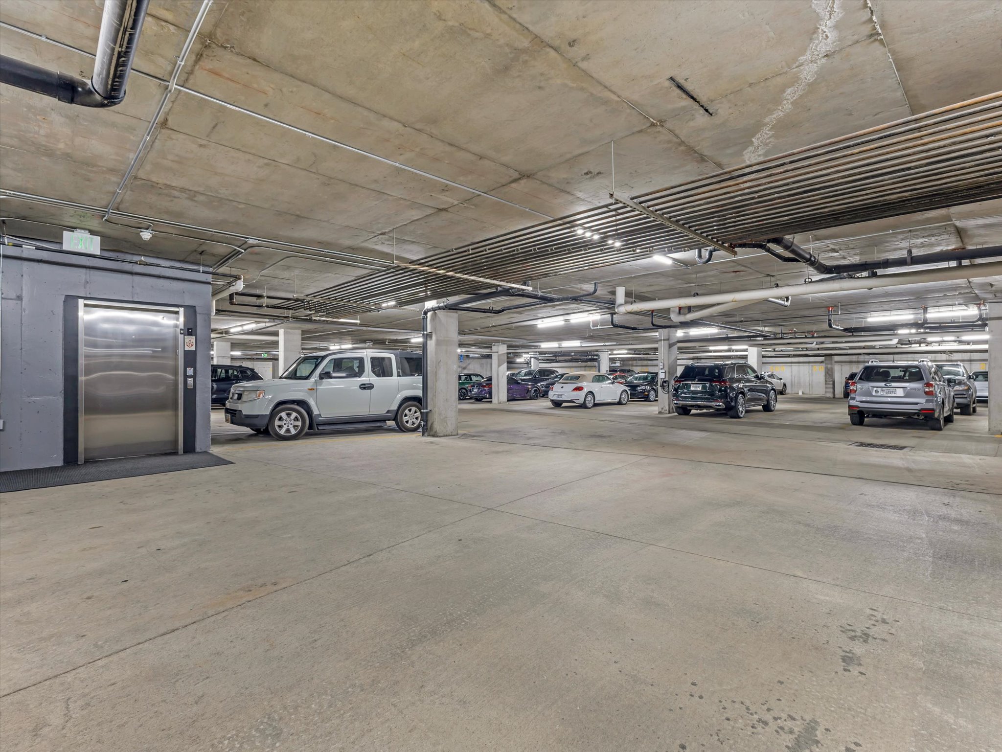 Underground parking garage featuring concrete flooring, concrete support columns, overhead lighting, exposed ceiling pipes, and a stainless steel elevator