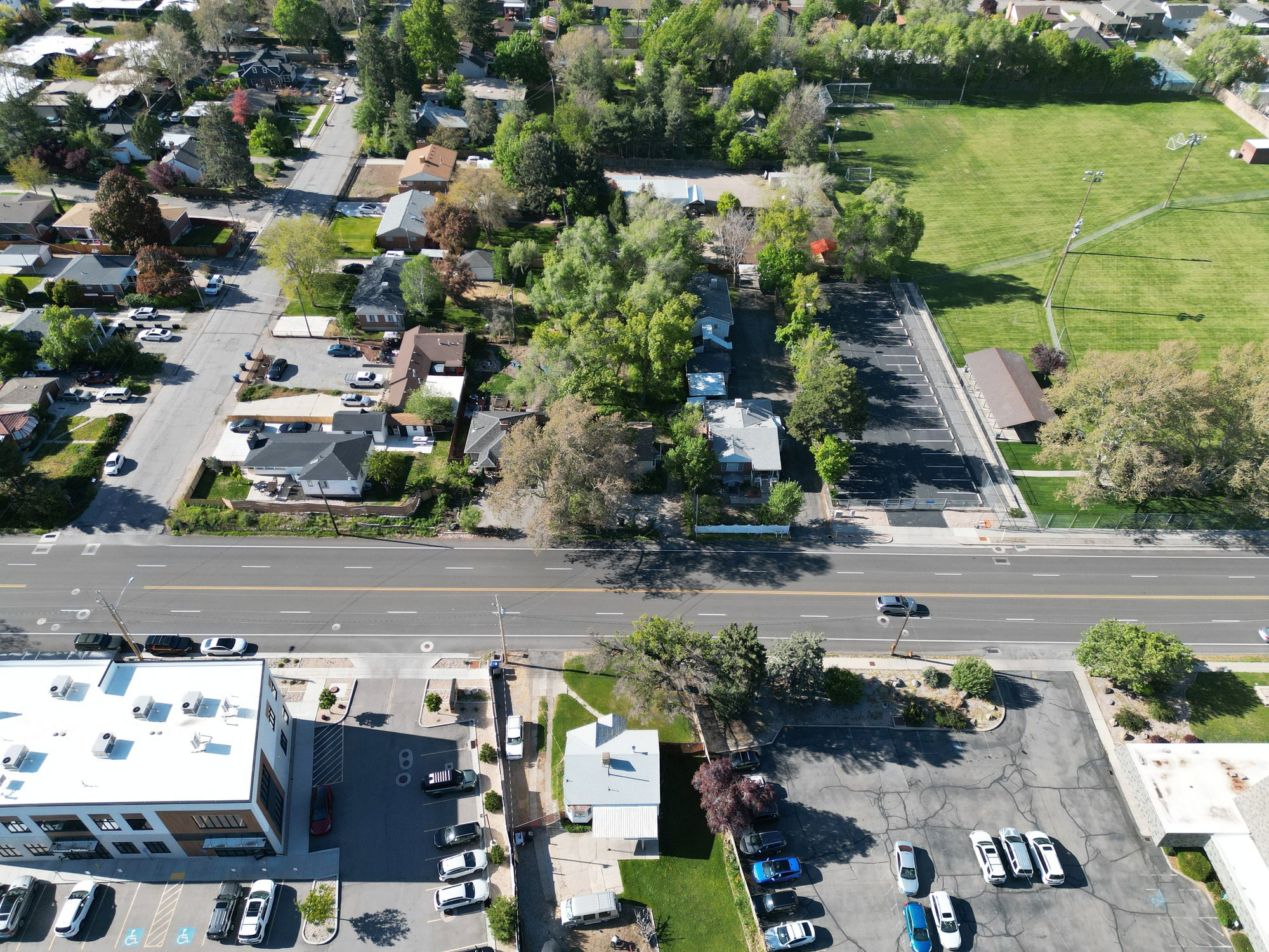 Urban aerial overview featuring a prominent multi-lane thoroughfare, surrounding residential properties with mature trees, commercial structures, and a sports field