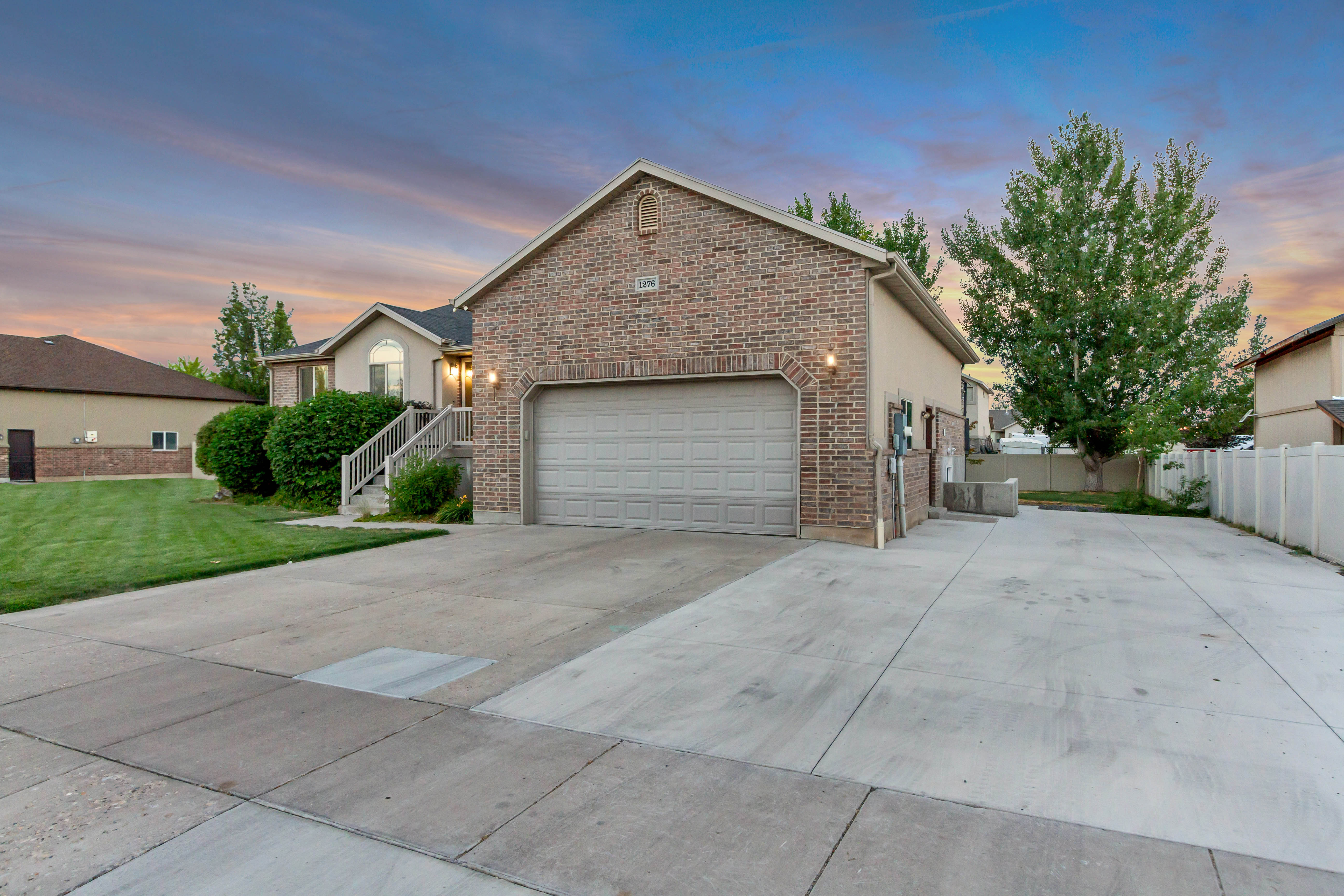 Brick facade property with an attached two-car garage, extended concrete driveway, mature landscaping, and a privacy fence
