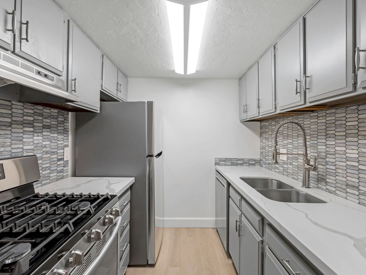 Modern galley kitchen featuring light gray cabinetry, stainless steel appliances, white patterned tile backsplash, light wood-finish flooring, and white countertops